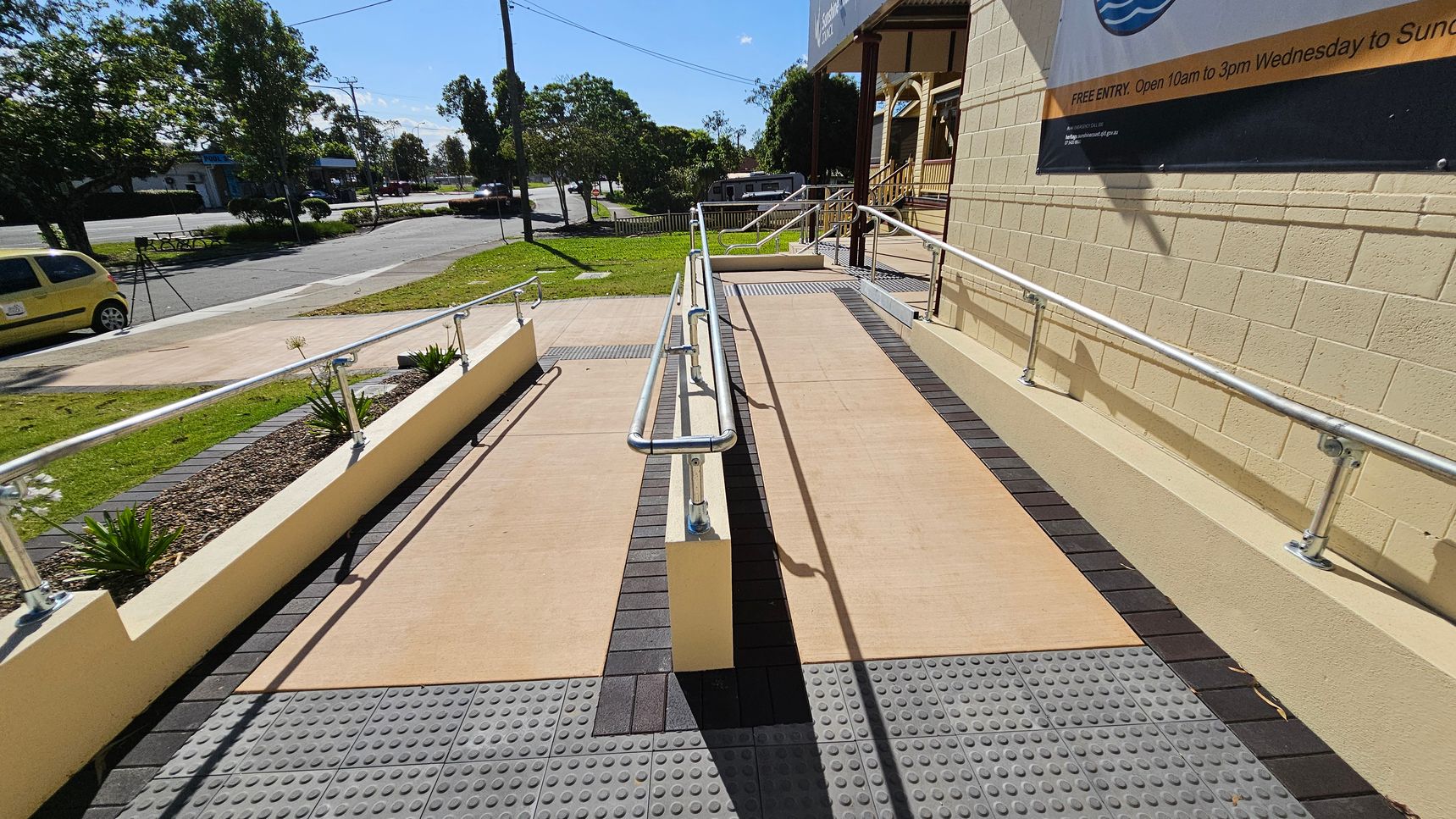 A concrete ramp with metal railings runs alongside a building with a beige brick wall on the right. The ramp is bordered by tactile paving. On the left, there's a small garden area with plants and a yellow car parked on the street. Trees and a petrol station are visible in the background. A large sign is mounted on the building wall. A concrete ramp with metal railings runs alongside a building with a beige brick wall on the right. The ramp is bordered by tactile paving. On the left, there's a small garden area with plants and a yellow car parked on the street. Trees and a petrol station are visible in the background. A large sign is mounted on the building wall.