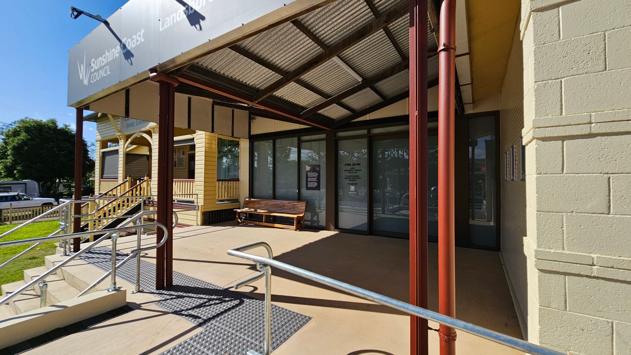 A building entrance with a covered porch supported by brown metal poles. The porch has a sloped roof with a metal frame and corrugated panels. A wooden bench is placed against the glass doors on the right. A ramp with metal railings leads up to the entrance. On the left, a yellow wooden structure with a small staircase and railing is visible. The ground is concrete, and there is a grassy area with a tree in the background. A building entrance with a covered porch supported by brown metal poles. The porch has a sloped roof with a metal frame and corrugated panels. A wooden bench is placed against the glass doors on the right. A ramp with metal railings leads up to the entrance. On the left, a yellow wooden structure with a small staircase and railing is visible. The ground is concrete, and there is a grassy area with a tree in the background.