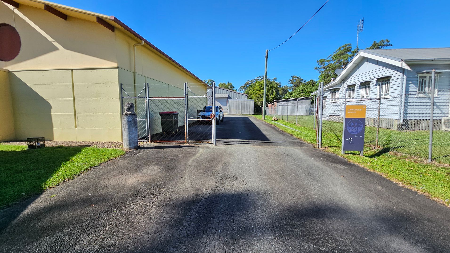 A narrow asphalt driveway leads to a fenced area with a gate. On the left, a large beige building casts a shadow on the grass. A white vehicle is parked inside the fenced area. On the right, a light blue building with multiple windows runs parallel to the driveway. A sign with text is positioned near the fence on the right side. Trees and a utility pole are visible in the background. A narrow asphalt driveway leads to a fenced area with a gate. On the left, a large beige building casts a shadow on the grass. A white vehicle is parked inside the fenced area. On the right, a light blue building with multiple windows runs parallel to the driveway. A sign with text is positioned near the fence on the right side. Trees and a utility pole are visible in the background.