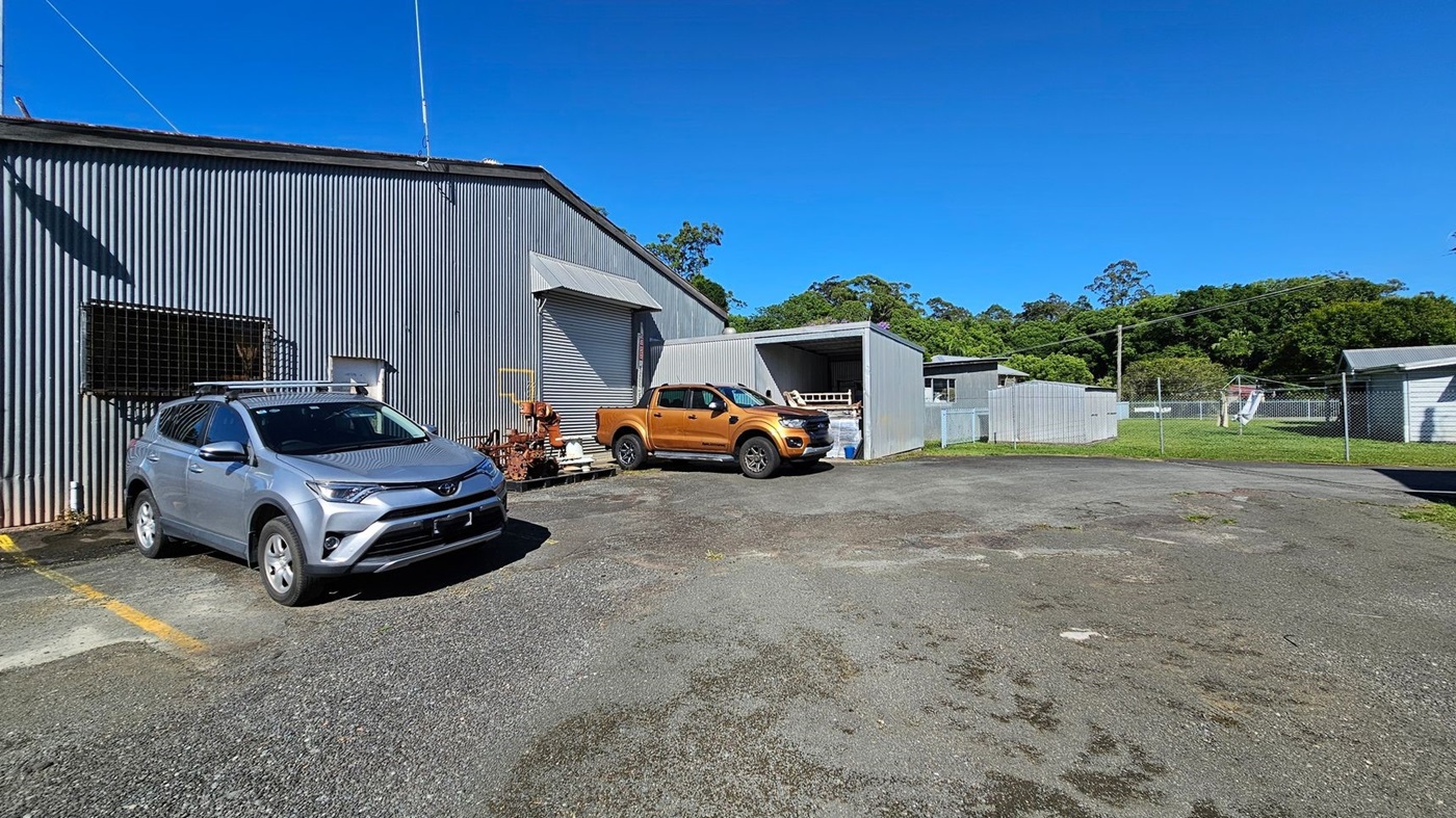 A corrugated metal building with a grey exterior is on the left, with a silver SUV parked in front. Next to it, a golden pickup truck is parked near a small structure with a roller door. The ground is a mix of gravel and patches of grass. In the background, there are more metal structures and a chain-link fence enclosing a grassy area with trees beyond. A corrugated metal building with a grey exterior is on the left, with a silver SUV parked in front. Next to it, a golden pickup truck is parked near a small structure with a roller door. The ground is a mix of gravel and patches of grass. In the background, there are more metal structures and a chain-link fence enclosing a grassy area with trees beyond.