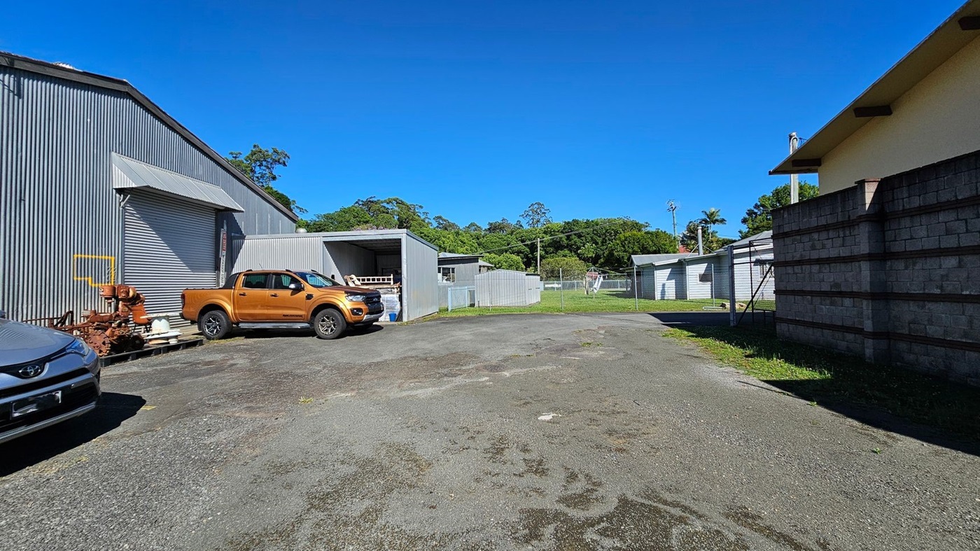A paved area with patches of grass and moss. On the left, there's a large corrugated metal building with a closed roller door, next to which is a rust-coloured pickup truck. A grey car is partially visible in the foreground. In the centre, a small open shed and a fenced grassy area are visible. On the right, a brick wall and a smaller building with a sloped roof. Trees and greenery are in the background. A paved area with patches of grass and moss. On the left, there's a large corrugated metal building with a closed roller door, next to which is a rust-coloured pickup truck. A grey car is partially visible in the foreground. In the centre, a small open shed and a fenced grassy area are visible. On the right, a brick wall and a smaller building with a sloped roof. Trees and greenery are in the background.