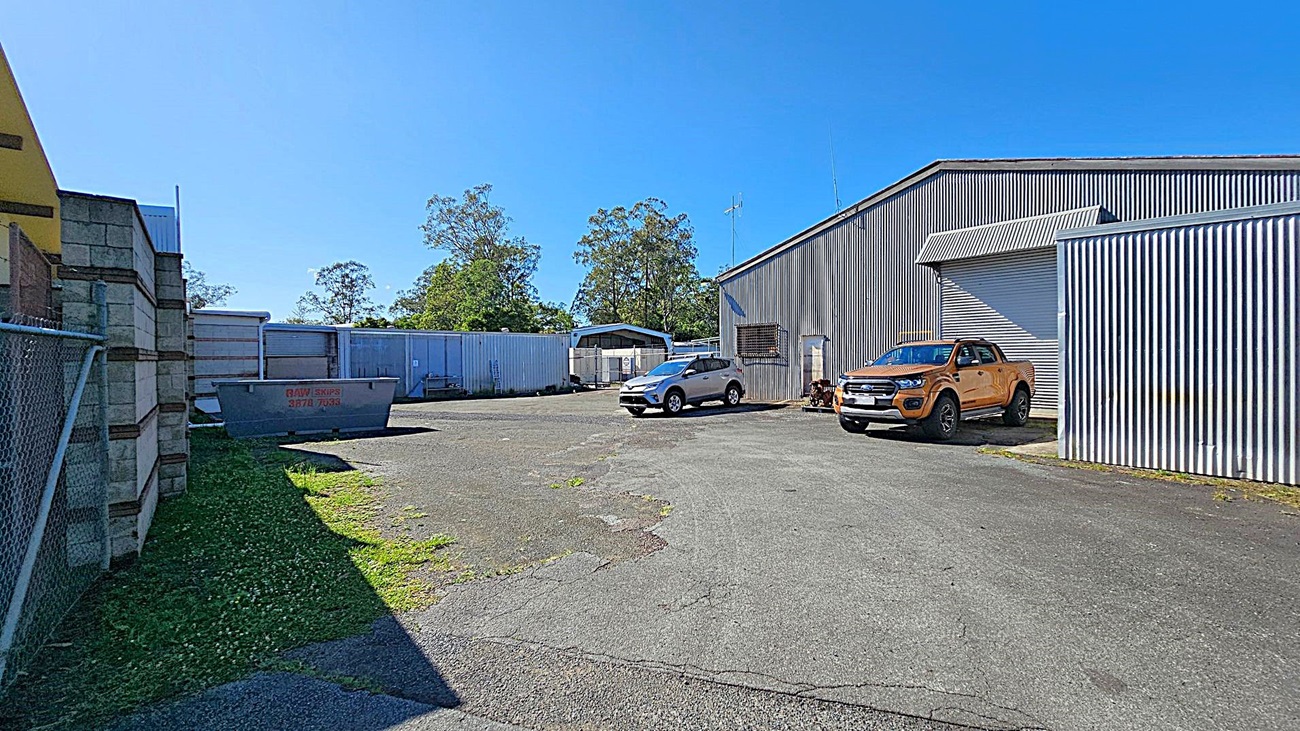 A paved industrial yard with a corrugated metal building on the right. Two vehicles, a silver SUV and an orange pickup truck, are parked in front of the building. On the left, a chain-link fence and a brick wall are visible. In the centre, a large metal skip bin is placed on the ground. In the background, there are more metal structures and some trees. The sky is clear and blue. A paved industrial yard with a corrugated metal building on the right. Two vehicles, a silver SUV and an orange pickup truck, are parked in front of the building. On the left, a chain-link fence and a brick wall are visible. In the centre, a large metal skip bin is placed on the ground. In the background, there are more metal structures and some trees. The sky is clear and blue.