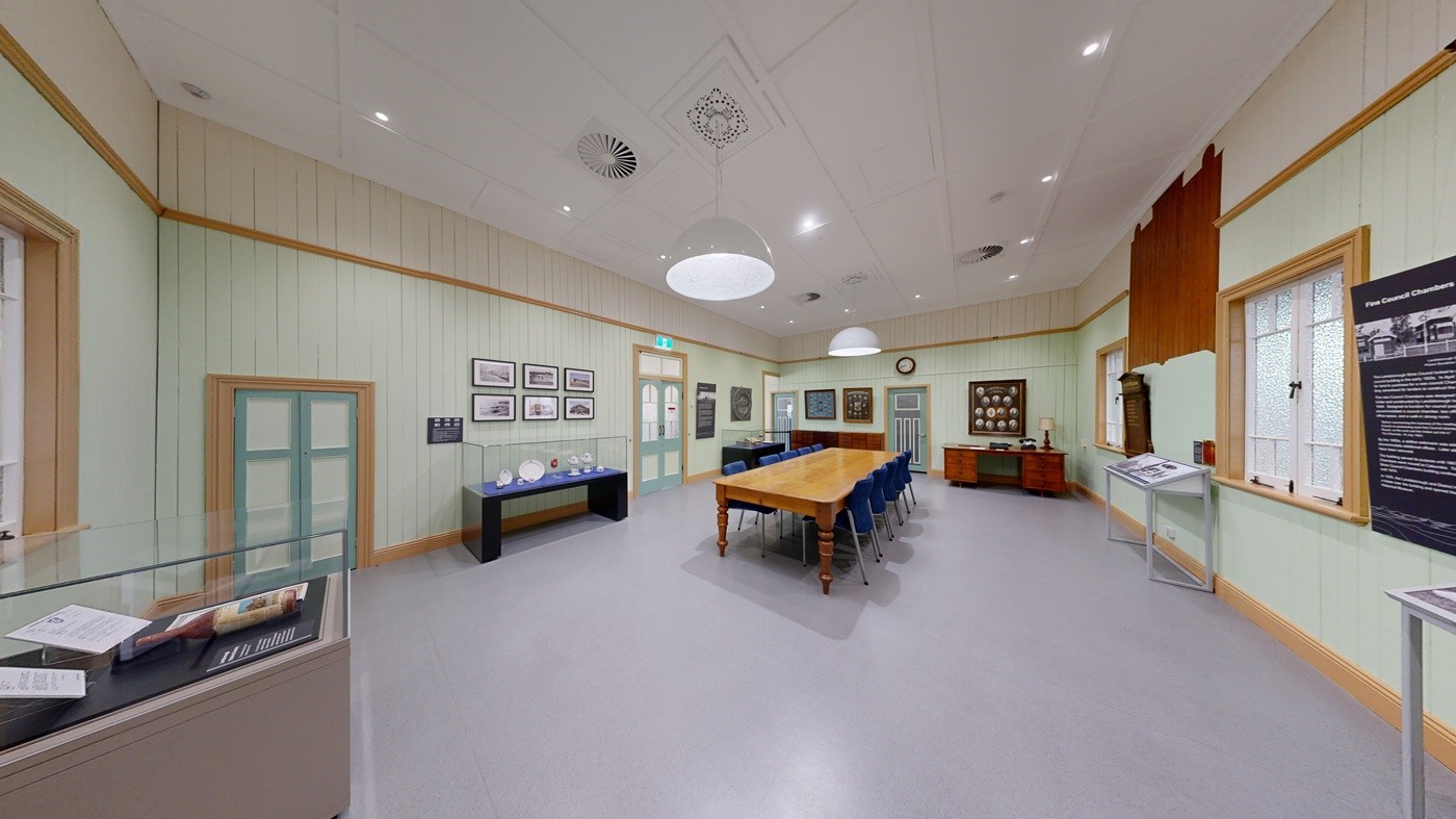 A spacious indoor area with a tiled floor and a wooden deck on the right, leading up to a building with cream-coloured weatherboard walls and large windows. The ceiling has exposed beams and metal trusses. On the left, there is a large frosted glass sliding door. Inside the building, tables and chairs are visible, and a sign marked "MACHINERY" points to the right. A small green pushchair is on the deck. A spacious indoor area with a tiled floor and a wooden deck on the right, leading up to a building with cream-coloured weatherboard walls and large windows. The ceiling has exposed beams and metal trusses. On the left, there is a large frosted glass sliding door. Inside the building, tables and chairs are visible, and a sign marked "MACHINERY" points to the right. A small green pushchair is on the deck.