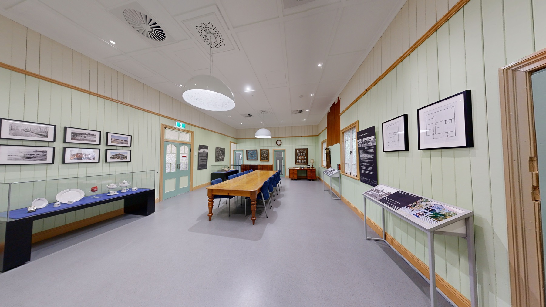 A room with a large wooden table surrounded by blue chairs on the left. The walls are light green with framed pictures and a large clock on the right. A door with a decorative design is also on the right. In the background, there are display cases with various items and a set of double doors. The ceiling has white panels with circular light fixtures and vents. The floor is light grey. A room with a large wooden table surrounded by blue chairs on the left. The walls are light green with framed pictures and a large clock on the right. A door with a decorative design is also on the right. In the background, there are display cases with various items and a set of double doors. The ceiling has white panels with circular light fixtures and vents. The floor is light grey.