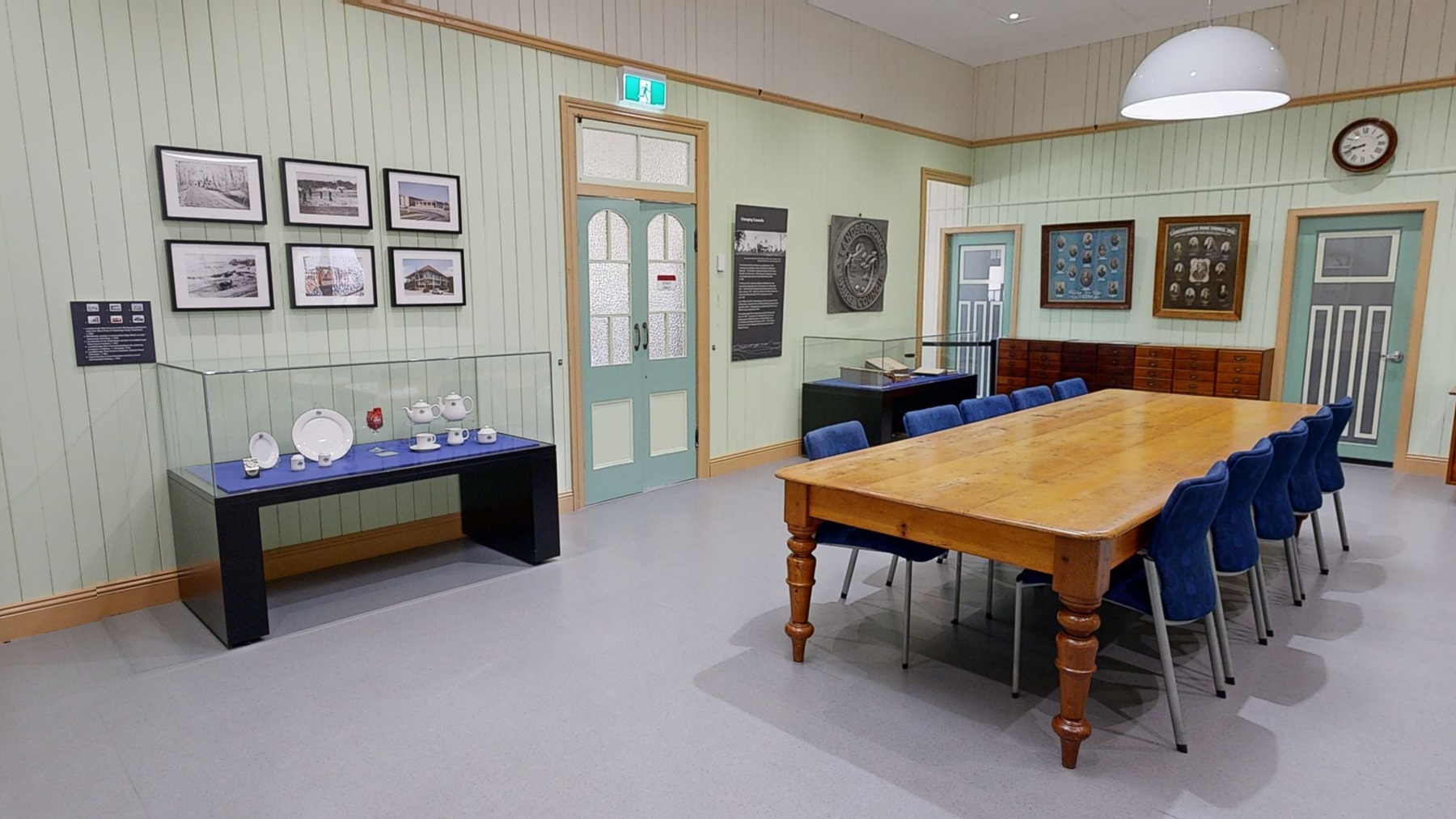 A room with light green panelled walls and a grey floor. On the left, framed photographs are displayed above a glass case containing white crockery on a blue surface. In the centre, a long wooden table with blue chairs runs parallel to the wall. The right side features a window, a wooden plaque, and informational displays. The ceiling is white with recessed lighting and a large round pendant light. A room with light green panelled walls and a grey floor. On the left, framed photographs are displayed above a glass case containing white crockery on a blue surface. In the centre, a long wooden table with blue chairs runs parallel to the wall. The right side features a window, a wooden plaque, and informational displays. The ceiling is white with recessed lighting and a large round pendant light.