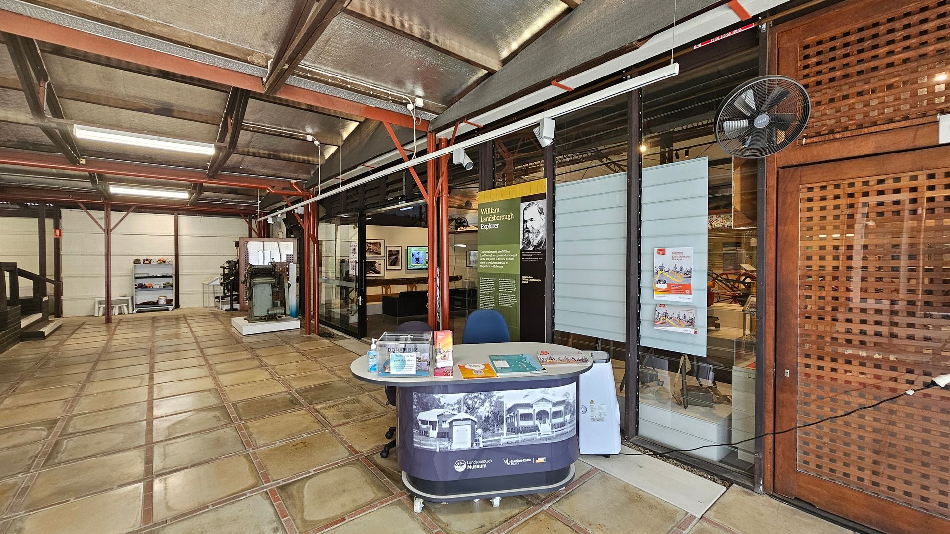 A museum interior with a tiled floor and a metal ceiling. On the right, there's a reception desk with brochures and a chair. Behind it, glass panels display information about William Landsborough. A wall-mounted fan is above. On the left, a display case with various items and a small table with stools are visible. The area is well-lit with fluorescent lights, and the structure features exposed beams. A museum interior with a tiled floor and a metal ceiling. On the right, there's a reception desk with brochures and a chair. Behind it, glass panels display information about William Landsborough. A wall-mounted fan is above. On the left, a display case with various items and a small table with stools are visible. The area is well-lit with fluorescent lights, and the structure features exposed beams.