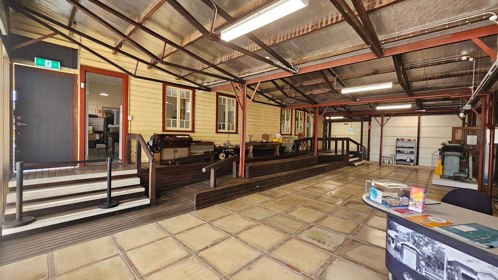 A spacious indoor area with a tiled floor and a corrugated metal ceiling. On the left, there is a black door with an exit sign above it, leading to a room with visible office furniture. Wooden steps and a railing lead up to a platform with vintage machinery and wooden benches. On the right, a curved table displays brochures and a clear box. Shelves with items are visible in the background, along with more machinery. The space is lit by fluorescent lights. A spacious indoor area with a tiled floor and a corrugated metal ceiling. On the left, there is a black door with an exit sign above it, leading to a room with visible office furniture. Wooden steps and a railing lead up to a platform with vintage machinery and wooden benches. On the right, a curved table displays brochures and a clear box. Shelves with items are visible in the background, along with more machinery. The space is lit by fluorescent lights.