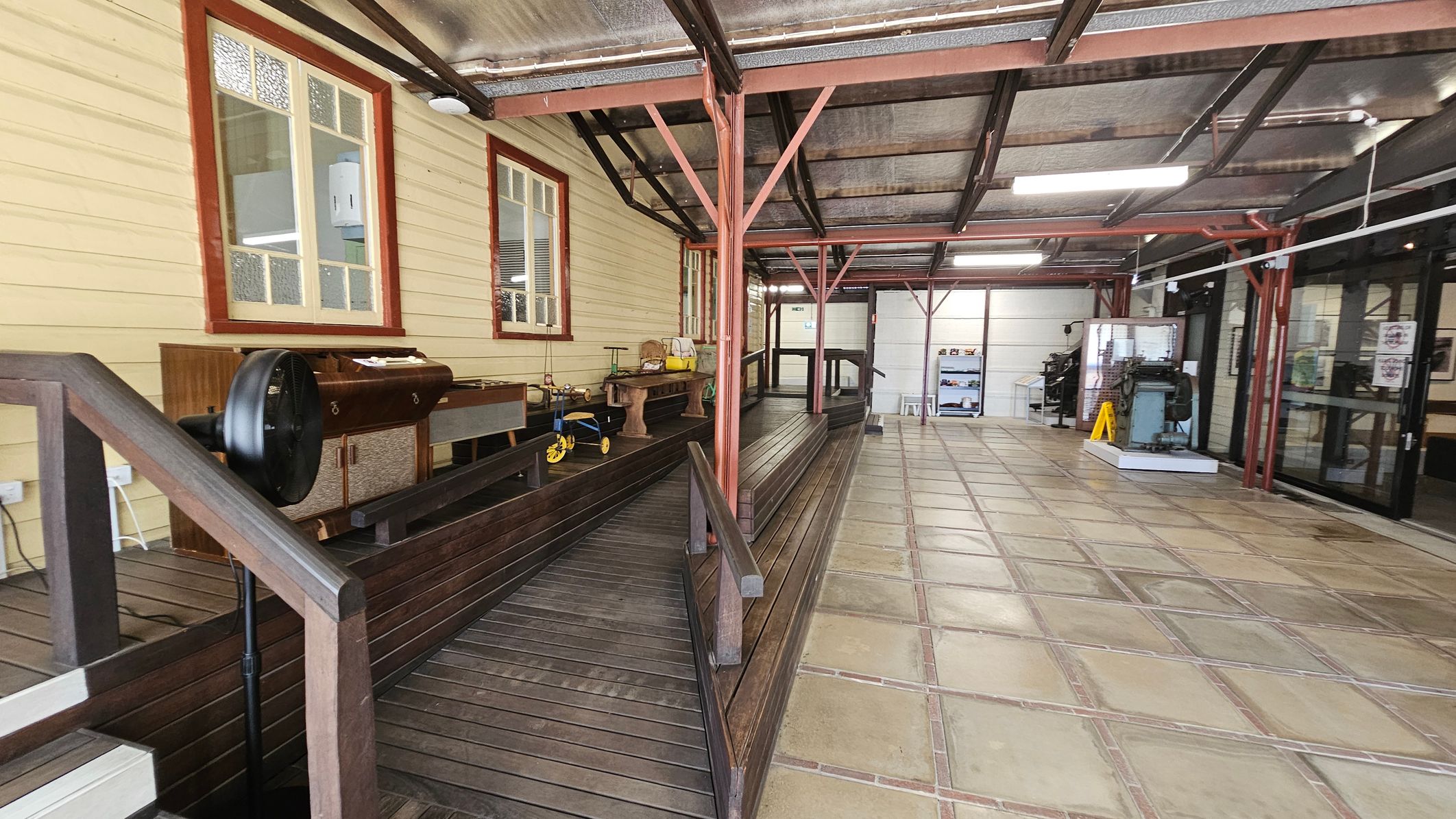 A covered walkway with a wooden ramp on the left, featuring a vintage tricycle and various items on a wooden bench. The wall has two windows with red frames. The right side has a tiled floor and a glass entrance with a yellow caution sign. The ceiling is metal with exposed beams and fluorescent lights. In the background, there is a display case and a machine on a platform. A covered walkway with a wooden ramp on the left, featuring a vintage tricycle and various items on a wooden bench. The wall has two windows with red frames. The right side has a tiled floor and a glass entrance with a yellow caution sign. The ceiling is metal with exposed beams and fluorescent lights. In the background, there is a display case and a machine on a platform.