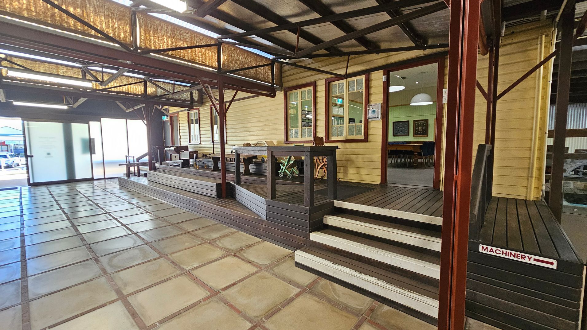 A spacious indoor area with a tiled floor and a wooden decked platform on the right. The platform has steps leading up to a building with cream-coloured wooden walls and large windows. Inside, there are tables and chairs visible. The ceiling has exposed beams and lighting fixtures. On the left, there is a large frosted glass sliding door, and outside, a glimpse of parked cars. A sign on the steps reads "MACHINERY" with an arrow pointing right. A spacious indoor area with a tiled floor and a wooden decked platform on the right. The platform has steps leading up to a building with cream-coloured wooden walls and large windows. Inside, there are tables and chairs visible. The ceiling has exposed beams and lighting fixtures. On the left, there is a large frosted glass sliding door, and outside, a glimpse of parked cars. A sign on the steps reads "MACHINERY" with an arrow pointing right.