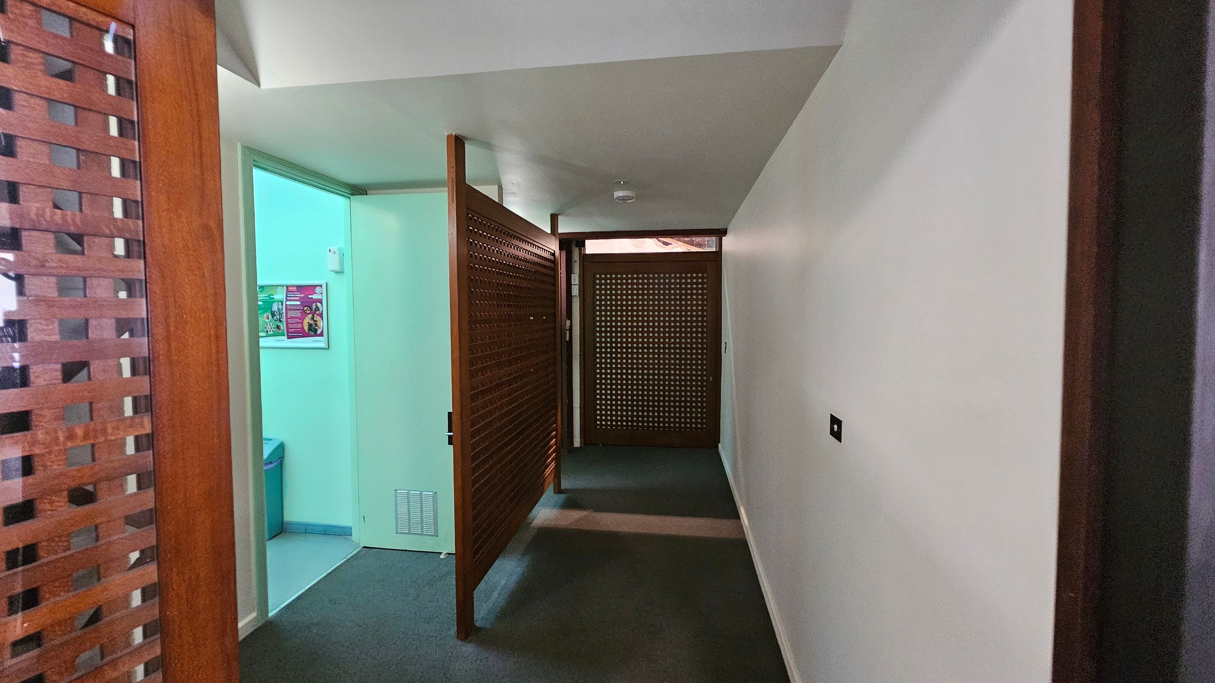 A hallway with a grey carpeted floor and white walls. On the left, there's a partially open wooden lattice door leading to a room with a greenish light. Inside, a poster is visible on the wall. Further down the hallway, another wooden lattice door is closed. The ceiling is plain white, and there is a small smoke detector mounted on it. A hallway with a grey carpeted floor and white walls. On the left, there's a partially open wooden lattice door leading to a room with a greenish light. Inside, a poster is visible on the wall. Further down the hallway, another wooden lattice door is closed. The ceiling is plain white, and there is a small smoke detector mounted on it.