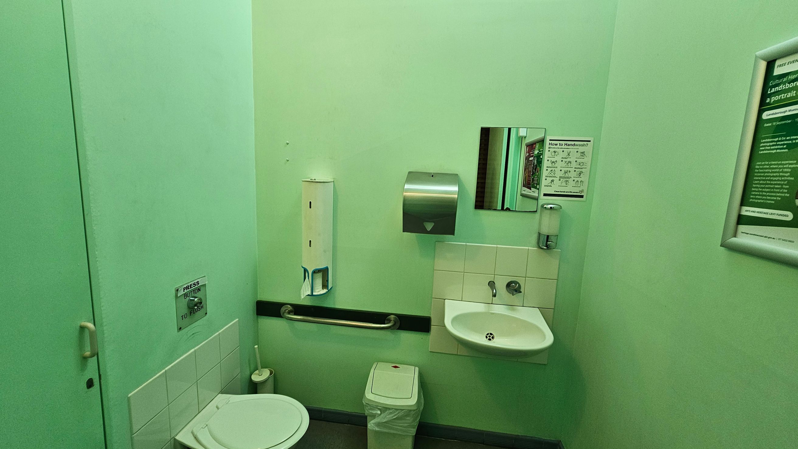 A small bathroom with light green walls. On the left, there's a toilet with a flush button above it and a toilet brush nearby. In the centre, a wall-mounted handrail, a paper towel dispenser, and a small mirror are visible. Below the mirror is a white sink with a soap dispenser. A waste bin is placed under the sink. On the right, there are framed informational posters. The floor is dark, and the ceiling is not visible. A small bathroom with light green walls. On the left, there's a toilet with a flush button above it and a toilet brush nearby. In the centre, a wall-mounted handrail, a paper towel dispenser, and a small mirror are visible. Below the mirror is a white sink with a soap dispenser. A waste bin is placed under the sink. On the right, there are framed informational posters. The floor is dark, and the ceiling is not visible.