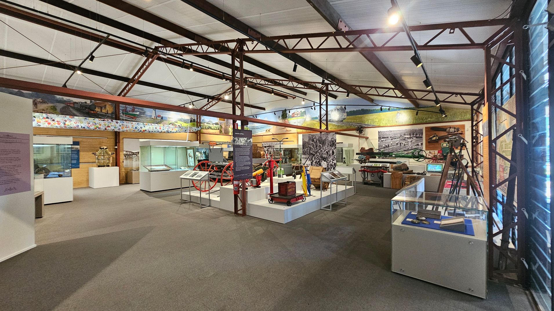 A museum interior with various exhibits displayed in glass cases and on platforms. On the left, there are informational panels and display cases. In the centre, historical machinery and artefacts are showcased, including a red wheel and other equipment. The right side features more display cases and a tripod. The ceiling has exposed metal beams and track lighting, and the floor is carpeted. Large murals and photographs decorate the walls. A museum interior with various exhibits displayed in glass cases and on platforms. On the left, there are informational panels and display cases. In the centre, historical machinery and artefacts are showcased, including a red wheel and other equipment. The right side features more display cases and a tripod. The ceiling has exposed metal beams and track lighting, and the floor is carpeted. Large murals and photographs decorate the walls.