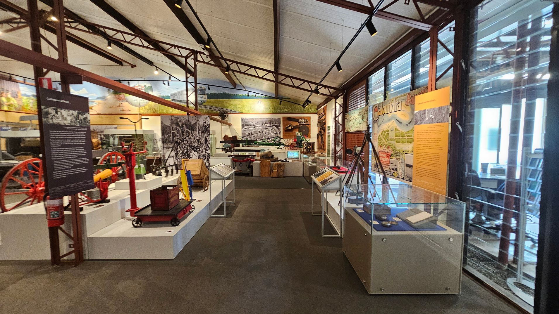 A museum interior with various exhibits. On the left, there are red and yellow machinery parts displayed on white platforms. In the centre, a large black-and-white photograph is mounted on the wall, with more machinery and artefacts below it. On the right, glass display cases contain documents and small items. The walls feature informational panels and historical images. The ceiling is supported by metal beams, and the floor is carpeted. Large windows on the right allow natural light in. A museum interior with various exhibits. On the left, there are red and yellow machinery parts displayed on white platforms. In the centre, a large black-and-white photograph is mounted on the wall, with more machinery and artefacts below it. On the right, glass display cases contain documents and small items. The walls feature informational panels and historical images. The ceiling is supported by metal beams, and the floor is carpeted. Large windows on the right allow natural light in.