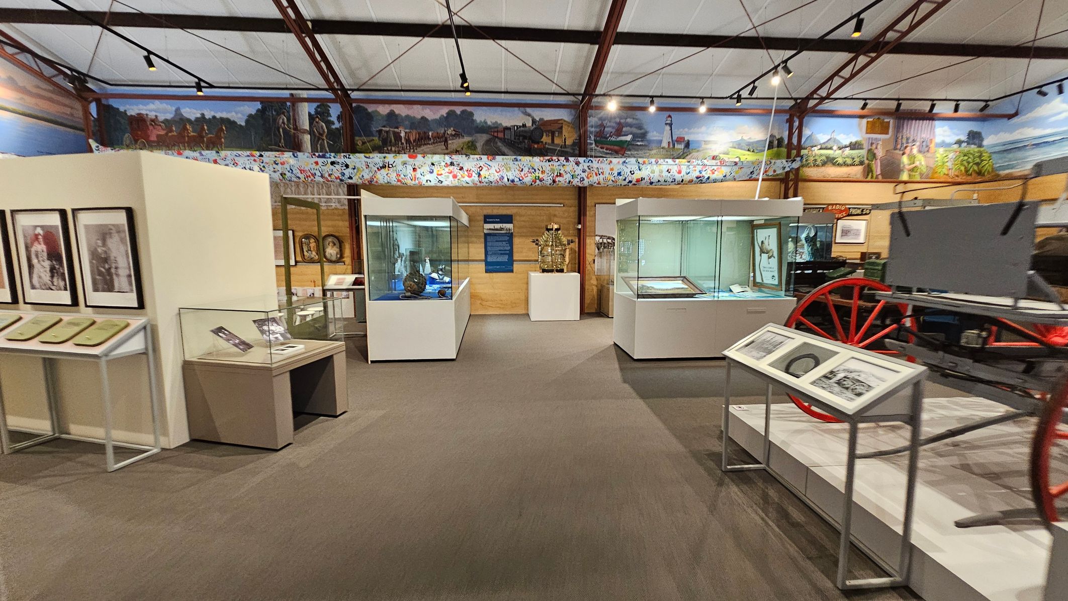 A museum interior with display cases and exhibits. On the left, there are framed photographs and documents on a stand. In the centre, glass display cases contain various artefacts, with a wooden wall and a blue information panel behind them. On the right, a red-wheeled carriage is partially visible, accompanied by informational panels. The ceiling is supported by metal beams, and a mural runs along the upper walls, depicting historical scenes. The floor is carpeted. A museum interior with display cases and exhibits. On the left, there are framed photographs and documents on a stand. In the centre, glass display cases contain various artefacts, with a wooden wall and a blue information panel behind them. On the right, a red-wheeled carriage is partially visible, accompanied by informational panels. The ceiling is supported by metal beams, and a mural runs along the upper walls, depicting historical scenes. The floor is carpeted.