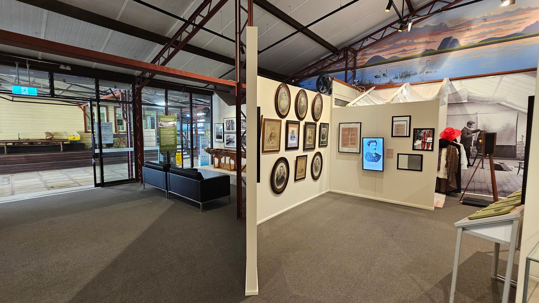 A museum interior with a carpeted floor and high ceiling. On the left, there's a glass entrance with a view of an outdoor area featuring a yellow building and wooden benches. In the centre, a wall displays various framed portraits and documents. To the right, there's a historical exhibit with vintage clothing and an old camera. A mural of a landscape with water and hills is visible above the exhibits. A small seating area with a black sofa is near the entrance. A museum interior with a carpeted floor and high ceiling. On the left, there's a glass entrance with a view of an outdoor area featuring a yellow building and wooden benches. In the centre, a wall displays various framed portraits and documents. To the right, there's a historical exhibit with vintage clothing and an old camera. A mural of a landscape with water and hills is visible above the exhibits. A small seating area with a black sofa is near the entrance.