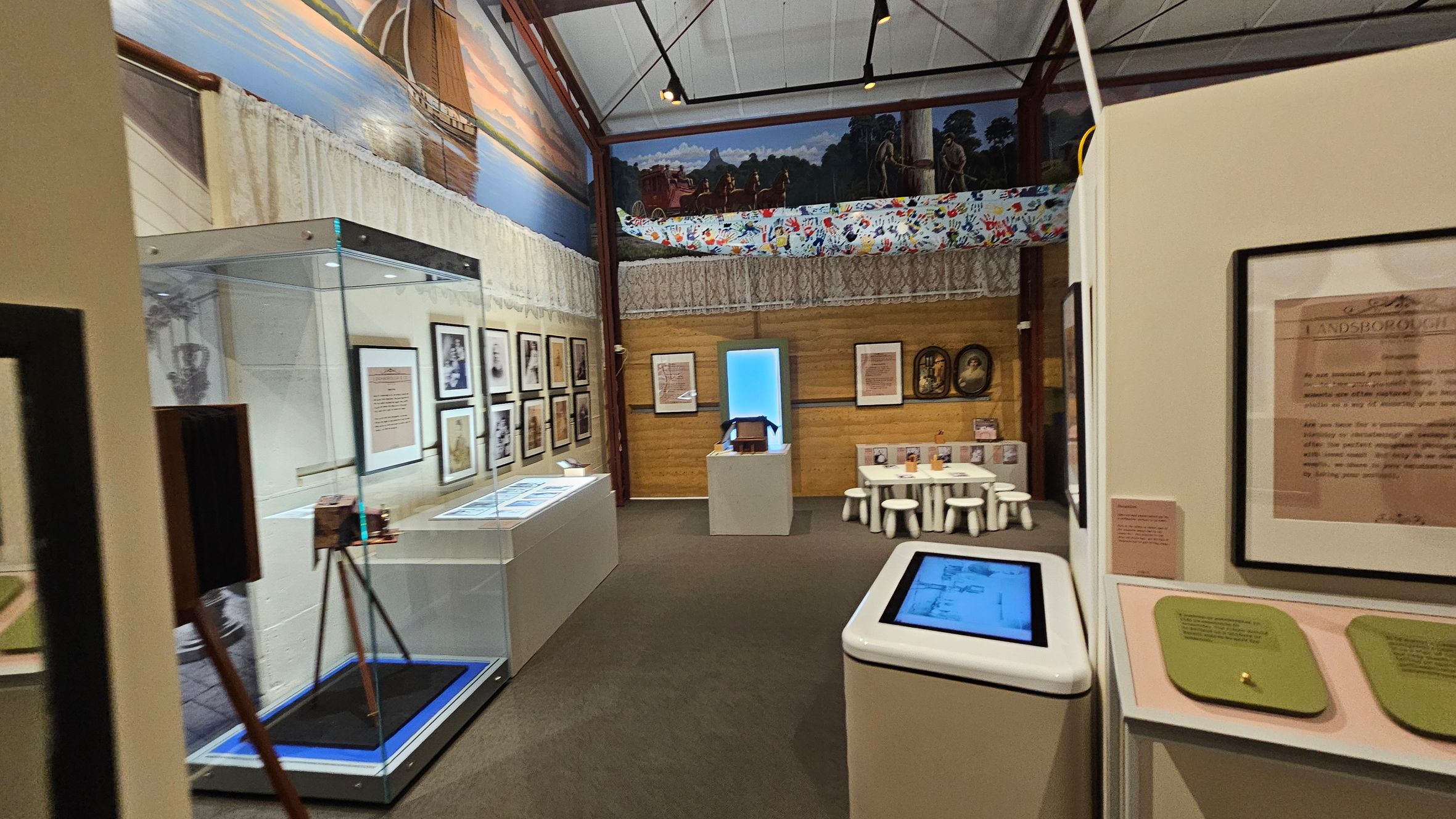 A museum exhibit with framed documents and photographs on the walls. On the left, a glass display case contains an object on a tripod. In the centre, a small table with white stools is set up, possibly for interactive activities. The background features a mural with a canoe and handprints. The ceiling is high with visible beams, and the floor is carpeted. An interactive screen is on the right. A museum exhibit with framed documents and photographs on the walls. On the left, a glass display case contains an object on a tripod. In the centre, a small table with white stools is set up, possibly for interactive activities. The background features a mural with a canoe and handprints. The ceiling is high with visible beams, and the floor is carpeted. An interactive screen is on the right.