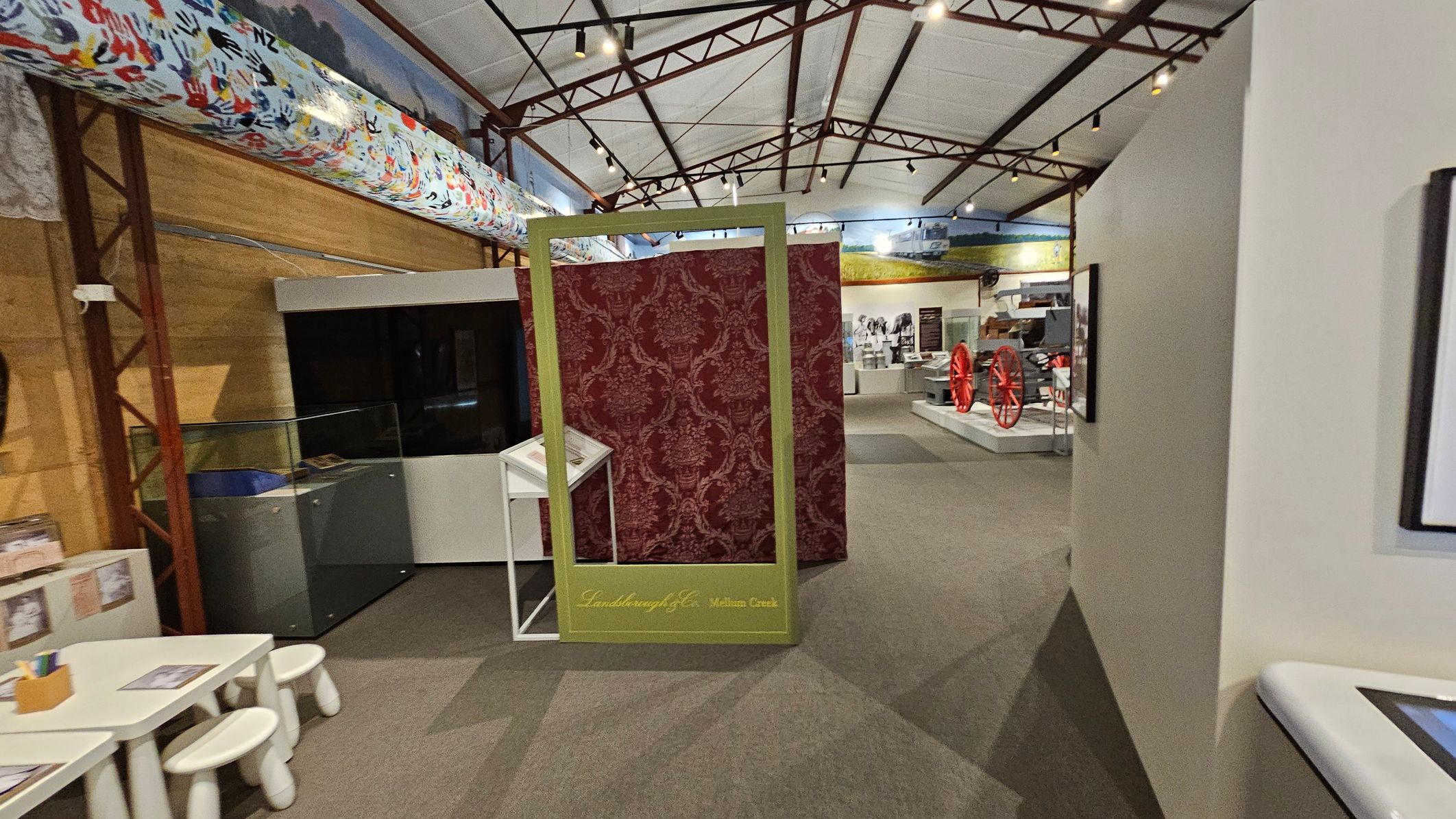 A museum interior with a high ceiling supported by metal beams. In the centre, a decorative red panel with a gold frame stands, labelled "Landsborough & Co. Melton Creek." To the left, a small table with stools and a display case. On the right, a wall with framed pictures. In the background, exhibits include red wagon wheels and historical artefacts. The floor is carpeted in a neutral tone. A museum interior with a high ceiling supported by metal beams. In the centre, a decorative red panel with a gold frame stands, labelled "Landsborough & Co. Melton Creek." To the left, a small table with stools and a display case. On the right, a wall with framed pictures. In the background, exhibits include red wagon wheels and historical artefacts. The floor is carpeted in a neutral tone.