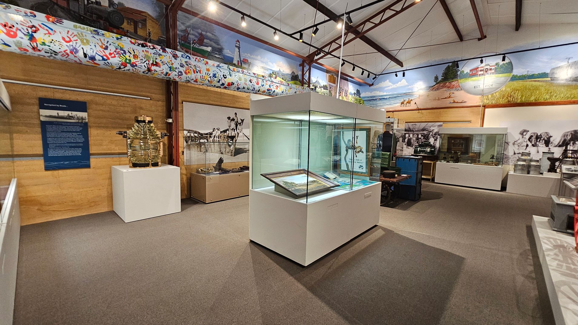 A museum interior with a central glass display case containing framed items. To the left, a large glass object on a white pedestal and a blue information panel are visible. The walls are adorned with murals depicting historical scenes and colourful handprints. The ceiling features exposed beams and track lighting. The floor is carpeted in grey. In the background, more display cases and historical photographs are present. A museum interior with a central glass display case containing framed items. To the left, a large glass object on a white pedestal and a blue information panel are visible. The walls are adorned with murals depicting historical scenes and colourful handprints. The ceiling features exposed beams and track lighting. The floor is carpeted in grey. In the background, more display cases and historical photographs are present.