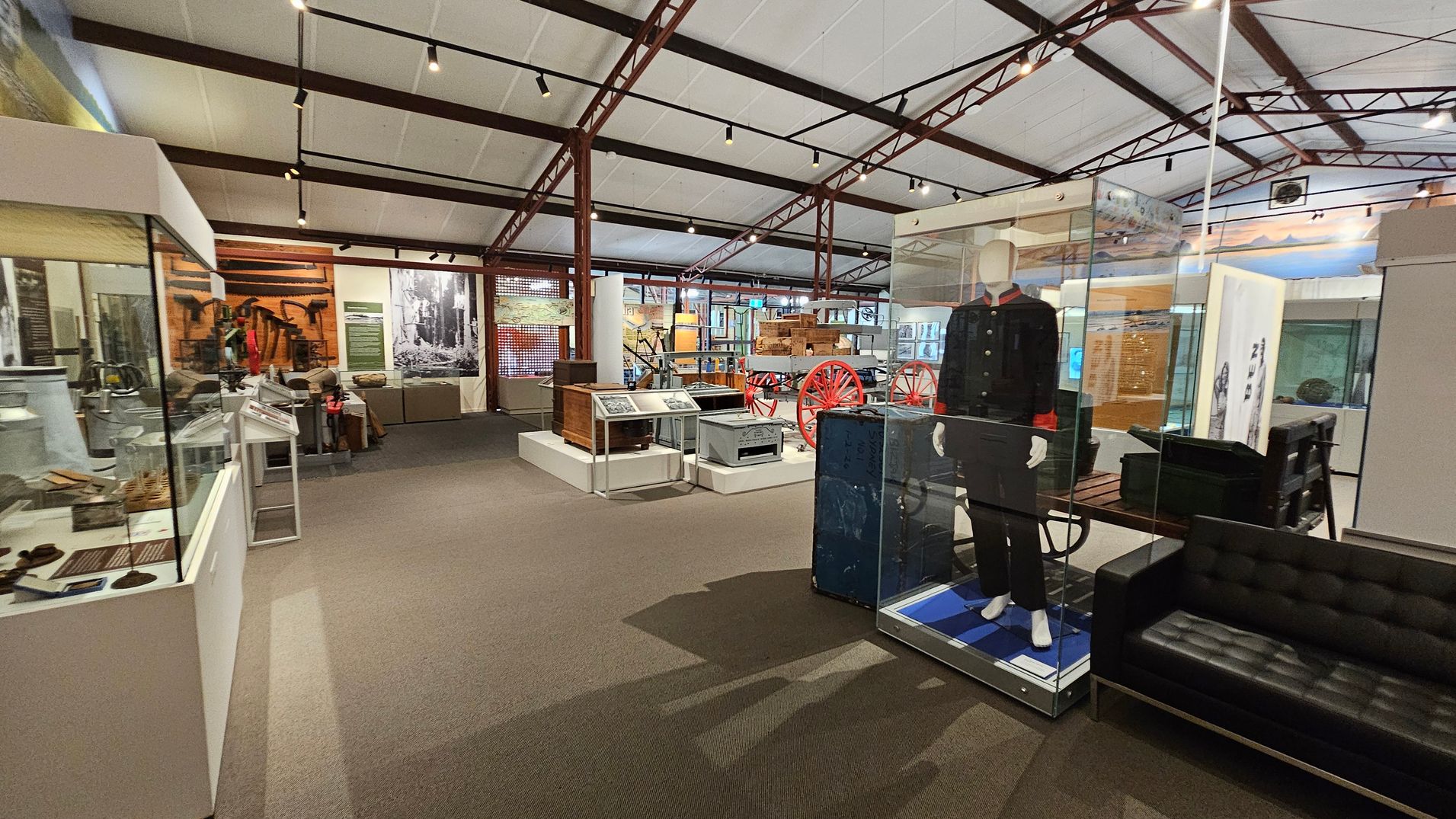 A museum interior with various exhibits. On the left, there are glass display cases containing artefacts. In the centre, a mannequin dressed in a historical uniform is encased in glass. Behind it, a red-wheeled cart is visible. The right side features a black leather sofa. The ceiling has exposed beams and track lighting, and the floor is carpeted. In the background, there are more displays and large black-and-white photographs on the walls. A museum interior with various exhibits. On the left, there are glass display cases containing artefacts. In the centre, a mannequin dressed in a historical uniform is encased in glass. Behind it, a red-wheeled cart is visible. The right side features a black leather sofa. The ceiling has exposed beams and track lighting, and the floor is carpeted. In the background, there are more displays and large black-and-white photographs on the walls.