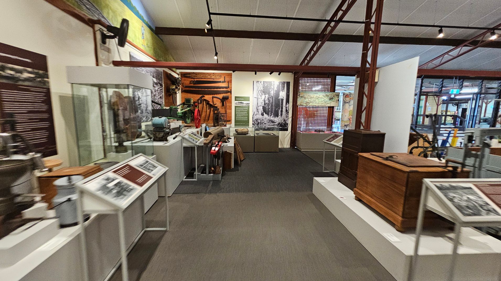 A museum exhibit with various historical artefacts. On the left, there are display cases with tools and informational panels. In the centre, a large black-and-white photograph is mounted on the wall, with more tools and equipment displayed below. On the right, wooden chests and additional artefacts are showcased. The floor is carpeted, and the ceiling has exposed beams with track lighting. A museum exhibit with various historical artefacts. On the left, there are display cases with tools and informational panels. In the centre, a large black-and-white photograph is mounted on the wall, with more tools and equipment displayed below. On the right, wooden chests and additional artefacts are showcased. The floor is carpeted, and the ceiling has exposed beams with track lighting.