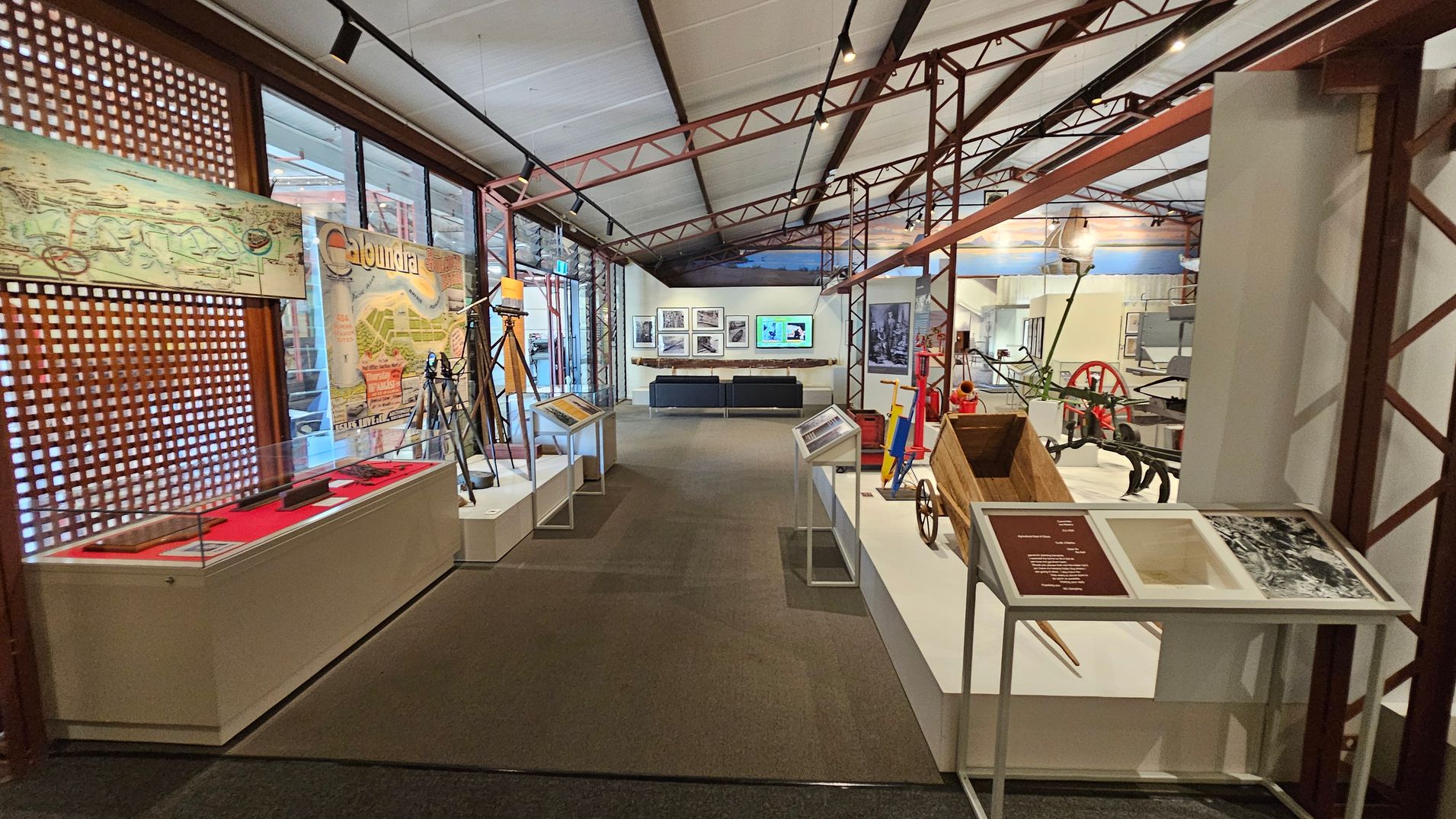 A museum exhibit with a high ceiling supported by metal beams. On the left, there's a display case with a red interior and a long object inside, alongside a wall with a large map and the word "Caloundra." In the centre, there are several tripods and information stands. On the right, a wooden wheelbarrow and other historical tools are displayed. The background features framed pictures and a screen above a black sofa. The floor is carpeted. A museum exhibit with a high ceiling supported by metal beams. On the left, there's a display case with a red interior and a long object inside, alongside a wall with a large map and the word "Caloundra." In the centre, there are several tripods and information stands. On the right, a wooden wheelbarrow and other historical tools are displayed. The background features framed pictures and a screen above a black sofa. The floor is carpeted.