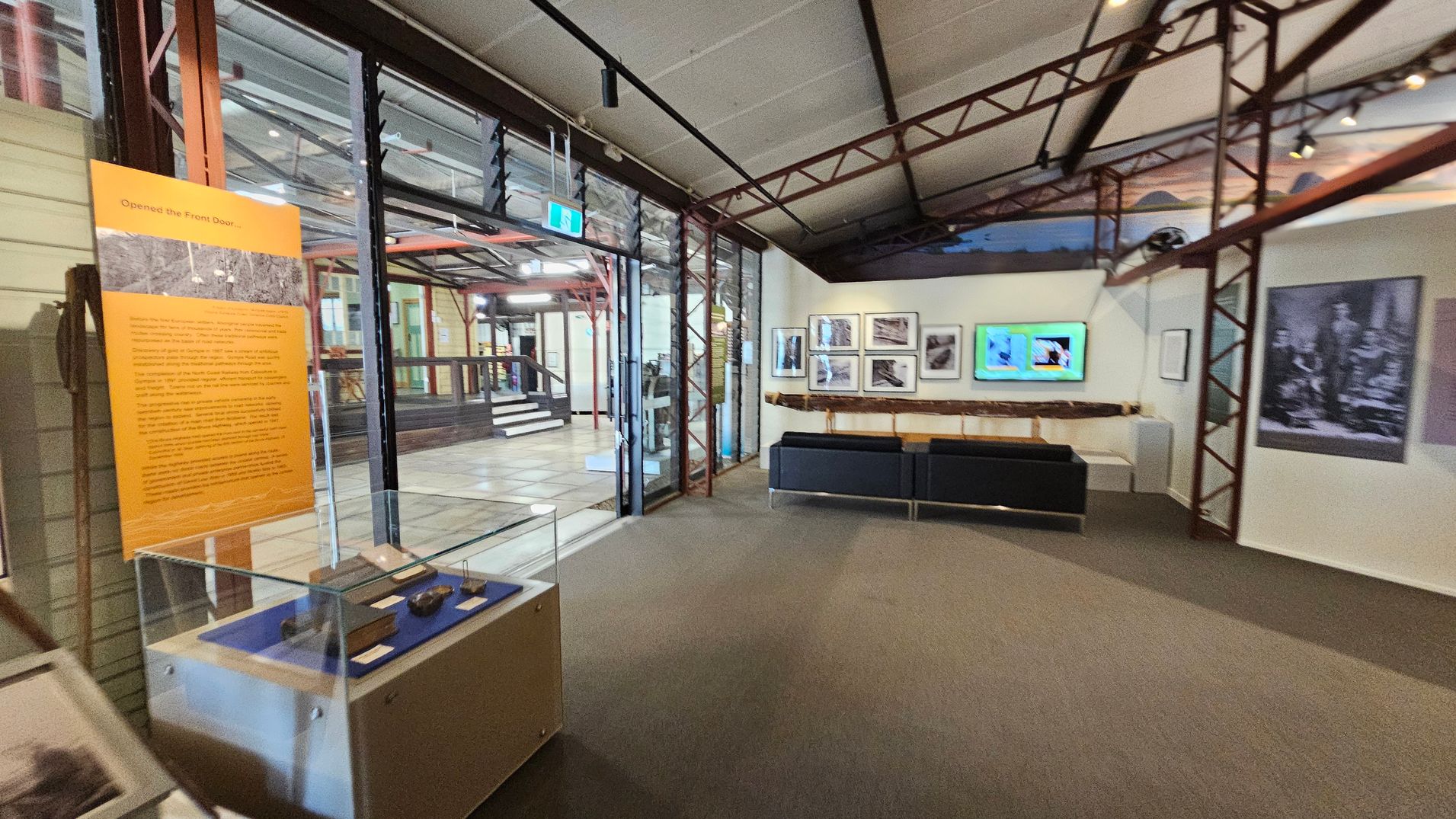 A museum interior with a glass display case on the left containing artefacts. An orange informational panel is nearby. The centre features a seating area with a black bench. On the right wall, there are framed photographs and a digital screen. The ceiling has exposed metal beams, and the floor is carpeted. In the background, a glass wall with a door leads to another section of the museum, visible through the glass. A museum interior with a glass display case on the left containing artefacts. An orange informational panel is nearby. The centre features a seating area with a black bench. On the right wall, there are framed photographs and a digital screen. The ceiling has exposed metal beams, and the floor is carpeted. In the background, a glass wall with a door leads to another section of the museum, visible through the glass.