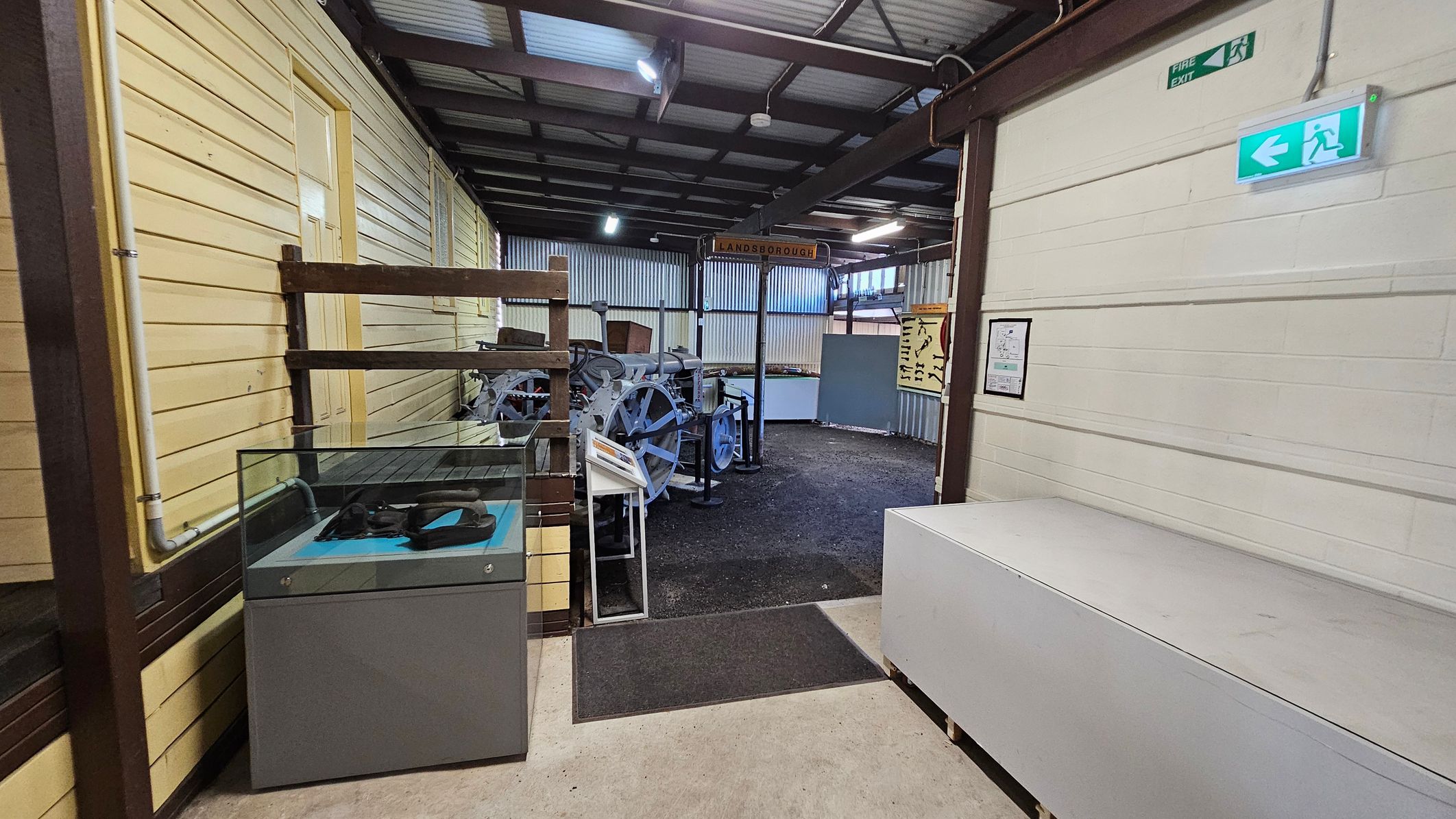 A room with a concrete floor and a corrugated metal ceiling. On the left, there's a wooden ramp with a "MACHINERY" sign. In the centre, a doorway leads to a space with machinery and display cases. On the right, a white brick wall features a small table with stools, colouring sheets, and markers. A shelf holds various items, including a green bag. A fire extinguisher is mounted on the wall near the doorway. A room with a concrete floor and a corrugated metal ceiling. On the left, there's a wooden ramp with a "MACHINERY" sign. In the centre, a doorway leads to a space with machinery and display cases. On the right, a white brick wall features a small table with stools, colouring sheets, and markers. A shelf holds various items, including a green bag. A fire extinguisher is mounted on the wall near the doorway.