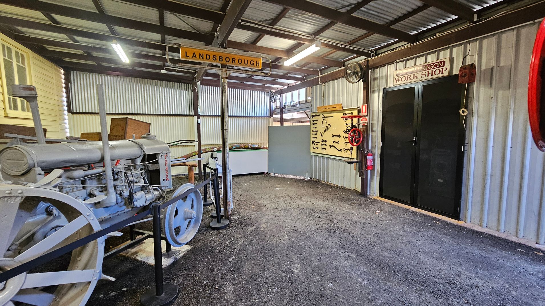 Inside a workshop with a corrugated metal ceiling and walls. On the left, there's a vintage Fordson tractor with large metal wheels. A sign above reads "LANDSBOROUGH." On the right, a black door labelled "WORK SHOP" is flanked by a red valve wheel and a fire extinguisher. A display board with various tools is mounted on the wall. The floor is gravel, and fluorescent lights hang from the ceiling. Inside a workshop with a corrugated metal ceiling and walls. On the left, there's a vintage Fordson tractor with large metal wheels. A sign above reads "LANDSBOROUGH." On the right, a black door labelled "WORK SHOP" is flanked by a red valve wheel and a fire extinguisher. A display board with various tools is mounted on the wall. The floor is gravel, and fluorescent lights hang from the ceiling.