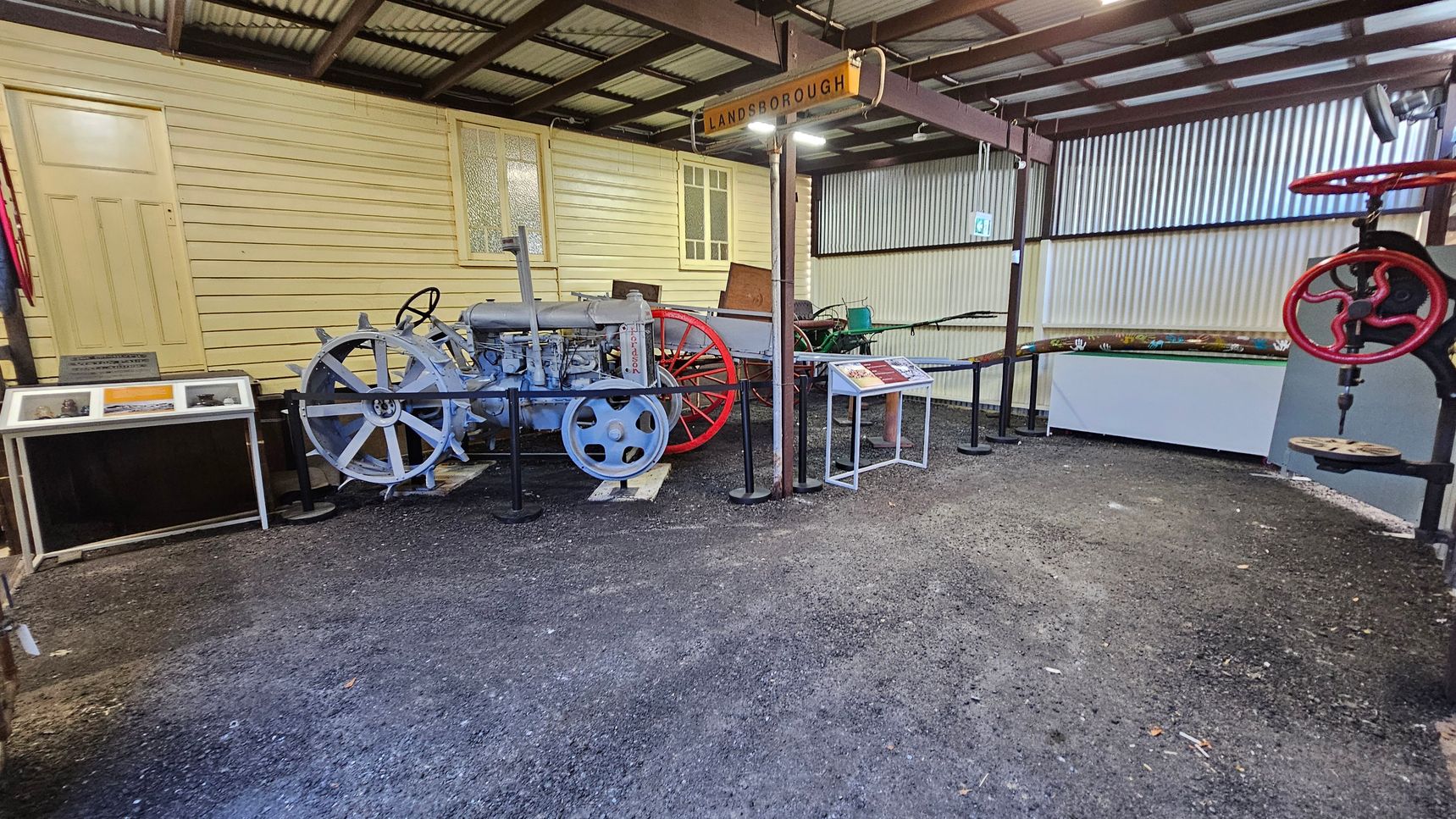 A museum exhibit featuring vintage machinery. On the left, there's a light blue tractor with large spoked wheels, displayed in front of a yellow wooden wall with a door and window. A sign above reads "LANDSBOROUGH." To the right, there's a red wheel mechanism and informational plaques. The floor is gravel, and the ceiling is corrugated metal. A museum exhibit featuring vintage machinery. On the left, there's a light blue tractor with large spoked wheels, displayed in front of a yellow wooden wall with a door and window. A sign above reads "LANDSBOROUGH." To the right, there's a red wheel mechanism and informational plaques. The floor is gravel, and the ceiling is corrugated metal.