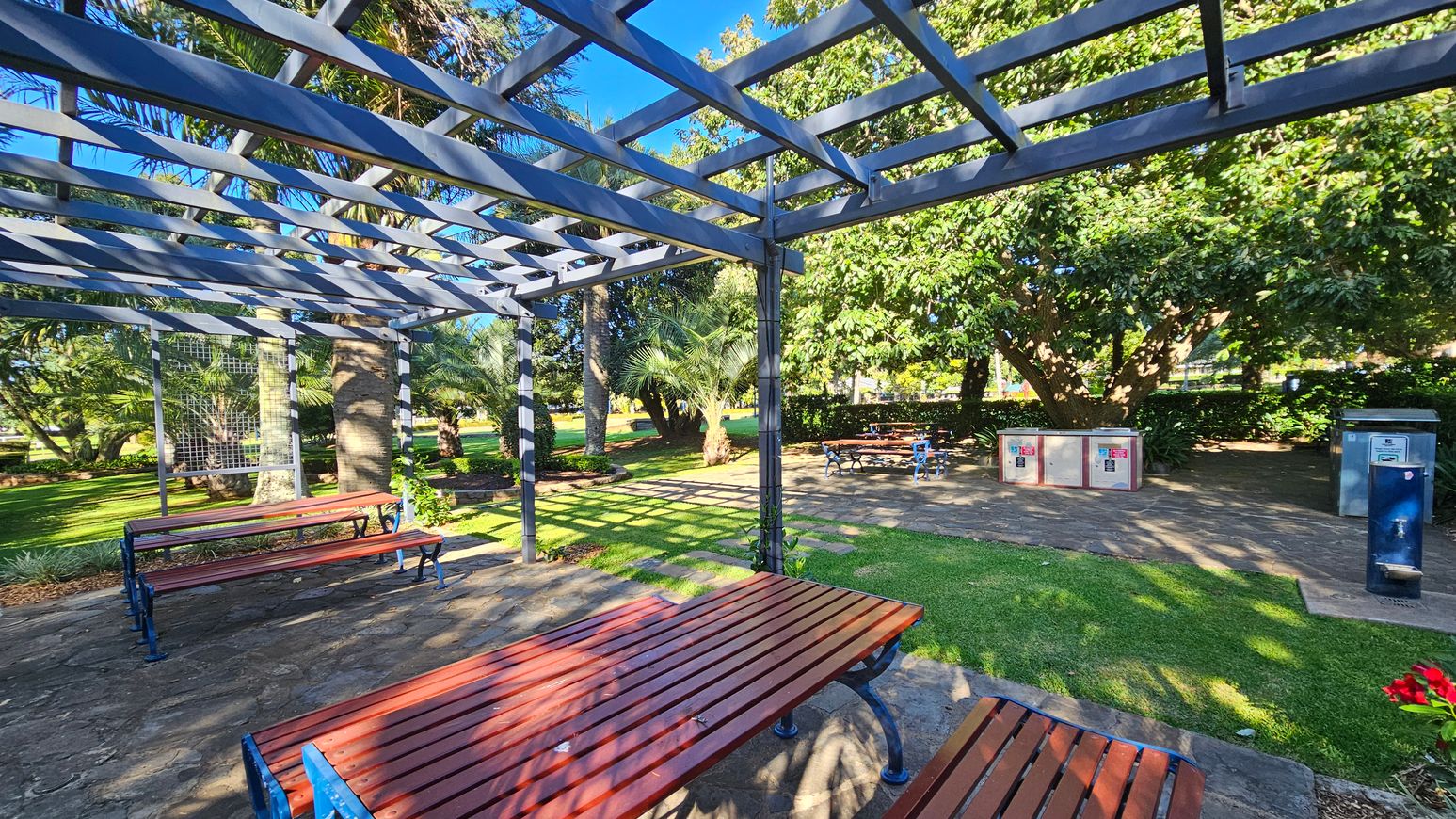 A park scene with a wooden picnic table and benches under a metal pergola in the foreground. The ground is a mix of stone paving and grass. In the background, there are more picnic tables, lush green trees, and a couple of utility boxes. A water fountain is visible on the right. Sunlight casts shadows through the pergola, creating patterns on the ground.