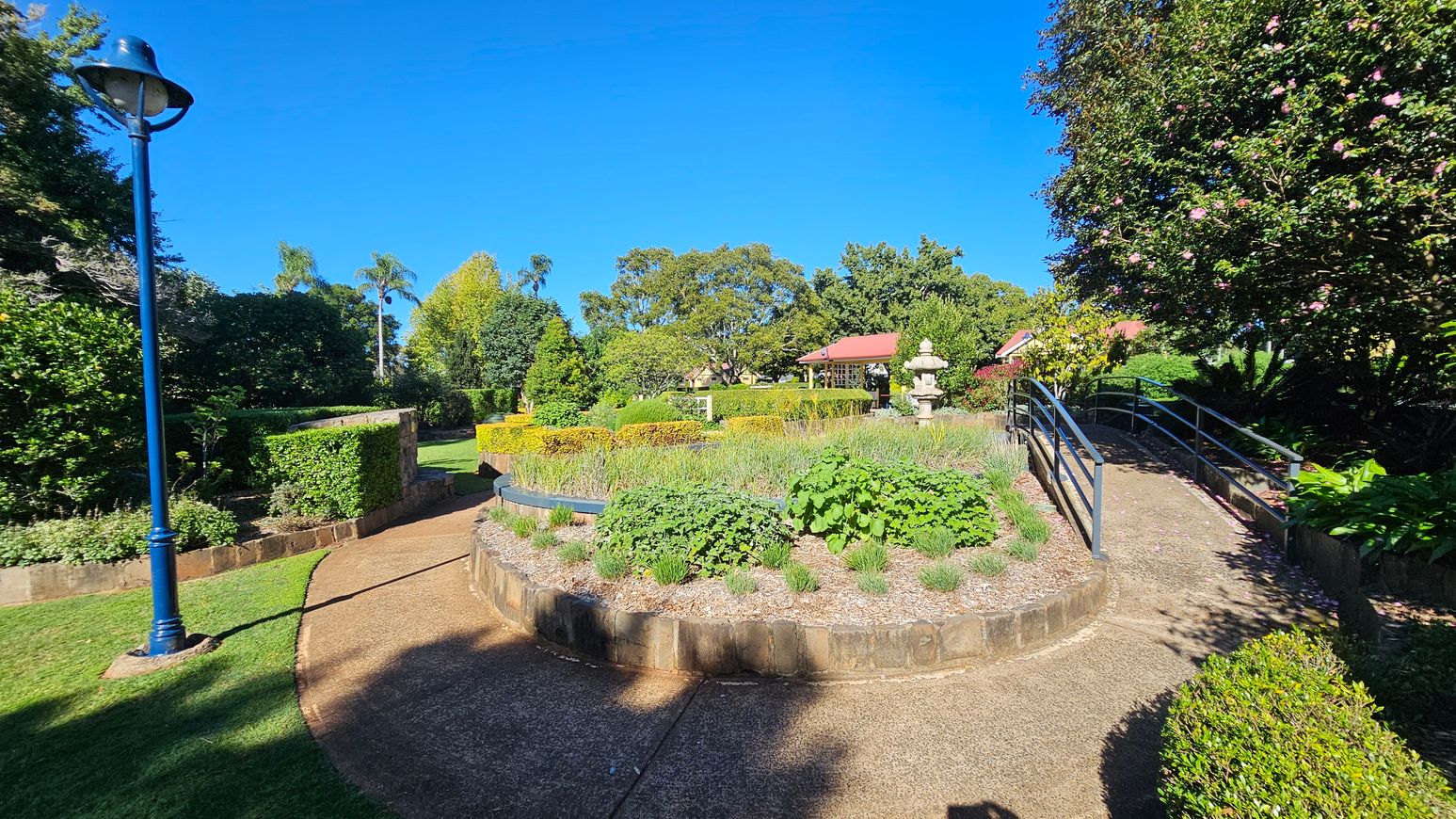 A garden scene with a curved path in the foreground, bordered by lush greenery and shrubs. On the left, a blue lamp post stands beside a hedge. In the centre, a raised circular garden bed is filled with various plants. To the right, a metal-railed ramp leads upwards, flanked by flowering bushes. In the background, a red-roofed structure and a stone lantern are partially visible among the trees.