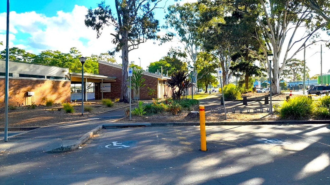 A closer look of the carpark area and access aisle designated for people with disability. Its background is the view of the large trees surrounding the open-air facility made of brown brick with a small reception area. A glimpse of the centre’s neighbourhood can be seen from afar.and a shade of white walls. The large trees surrounding the area provide shade due to its spreading canopy and crown.