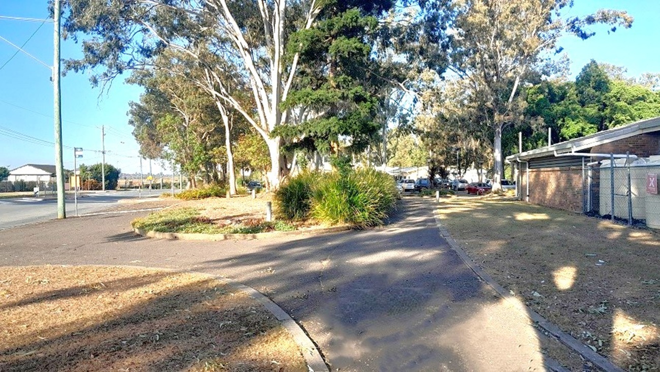 This image shows two accessible asphalted roads. The road to the left leads you to the main road and the other route will bring you to the carpark area. There are also huge trees surrounding the whole facility and part of the brown brick building with white gate. The wide-spreading canopy and crown of the large trees surrounding the building provides shade and a bright atmosphere.