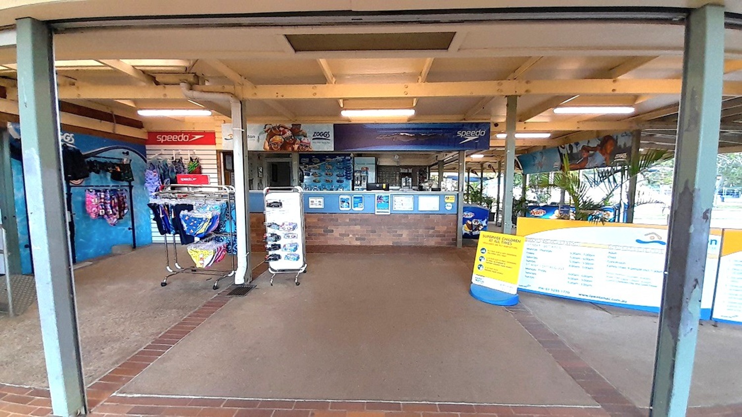 A shop interior with a tiled floor and a wooden ceiling. In the centre, there is a counter with various signs and brochures. To the left, racks display swimwear and accessories, with a "Speedo" sign above. On the right, large informational boards are visible, along with some potted plants. The shop is open to the outside, with a view of greenery in the background.