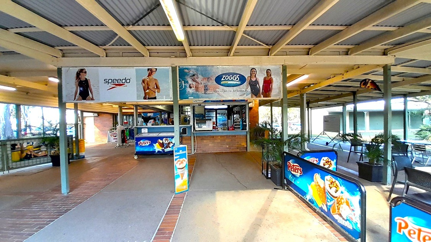 A covered outdoor area with a snack bar in the centre, featuring a counter with a drinks display. Above, there are advertisements for swimwear brands. On the left, there is a seating area with tables and chairs, partially enclosed by glass. On the right, more seating is visible, with tables and chairs under the roof. The floor is a mix of tiles and concrete, and the ceiling is made of corrugated metal with fluorescent lighting. Signs for ice cream are displayed prominently.