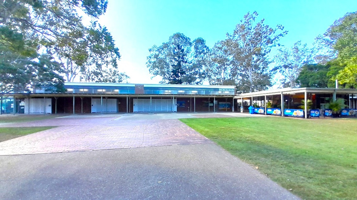 A single-storey building with a flat roof is in the centre, featuring a series of closed roller doors. To the right, there's an extended covered area with colourful signage and bins. The foreground has a paved path leading to the building, flanked by grass. In the background, tall trees with dense foliage are visible, providing a natural backdrop. The left side has more trees, adding to the greenery surrounding the structure.