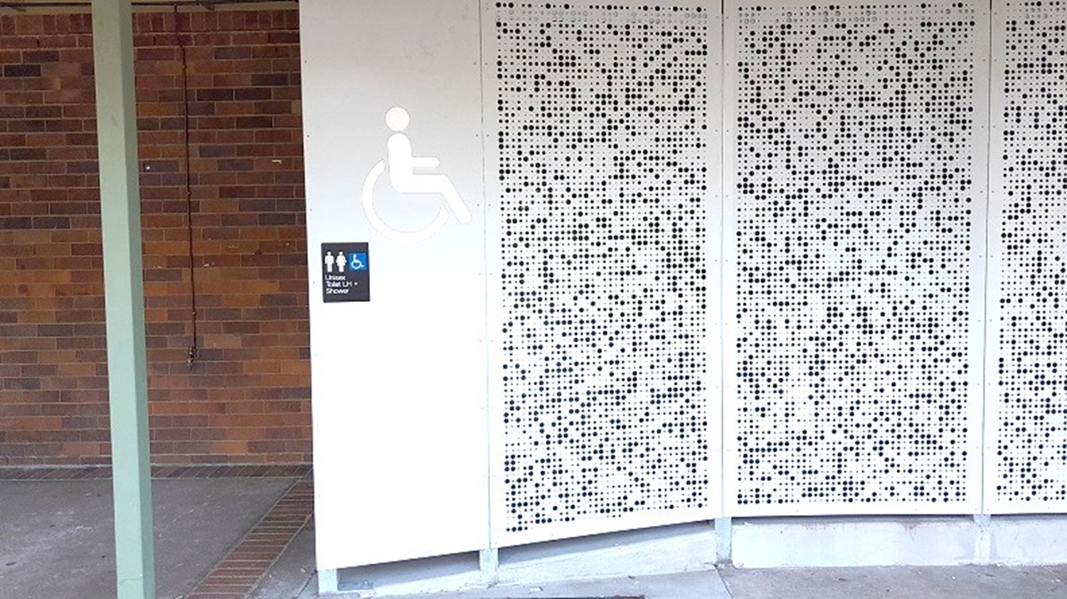 A wall with a perforated metal panel on the right, featuring a pattern of small, evenly spaced holes. To the left, a sign indicates accessible toilets, with symbols for male, female, and wheelchair access. The background has a brick wall partially visible. The floor is concrete, and a vertical green beam is on the far left.
