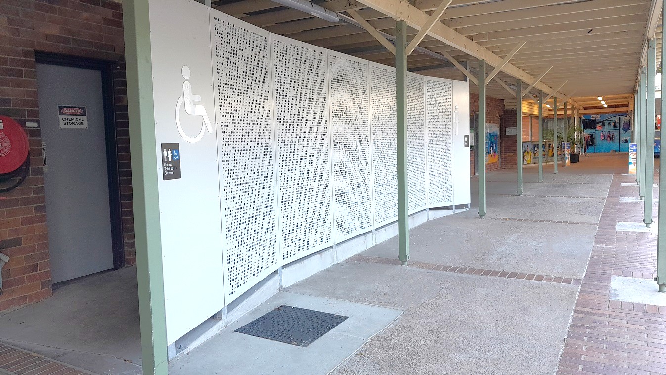 On the left, a door with a sign reading "Accessible Toilet" is set into a brick wall. Next to it, a white panel with a wheelchair symbol indicates accessibility. The middle features a curved, perforated white screen with a pattern of small holes. The right side shows a covered walkway with a wooden ceiling and green metal supports. The floor is a mix of concrete and brick, leading to a more open area in the background.