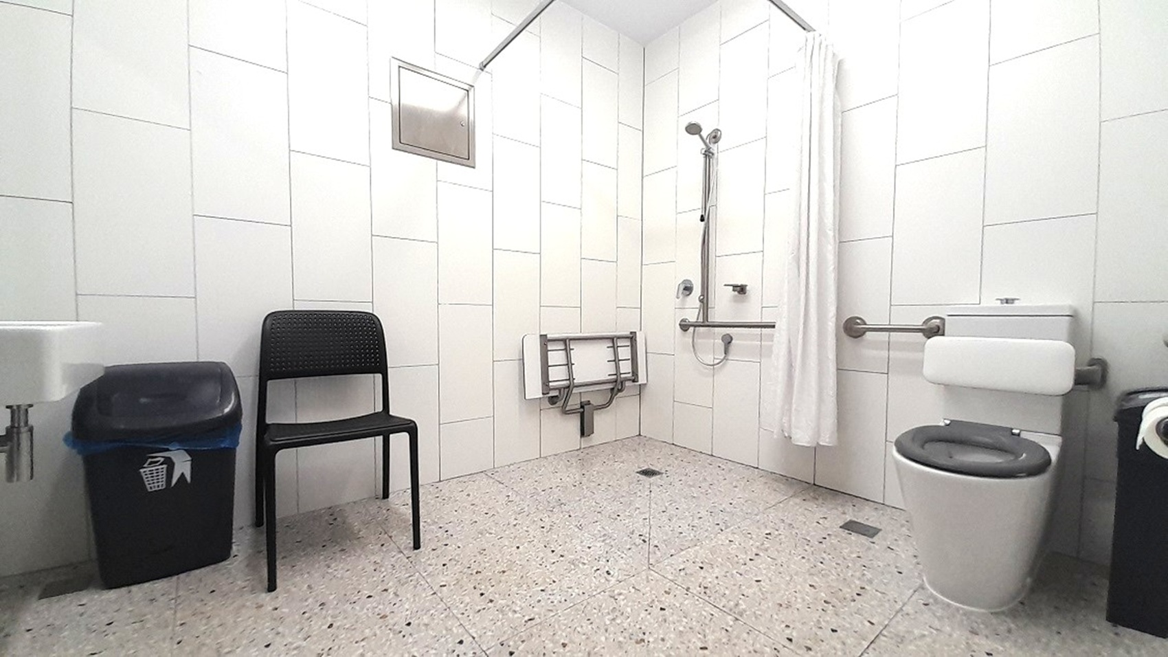 A bathroom with white tiled walls and a speckled floor. On the left, there's a black chair and a waste bin. In the centre, a shower area with a handheld showerhead, a foldable seat, and a curtain. On the right, a toilet with grab bars and a toilet paper holder. A small window is near the ceiling on the left wall.