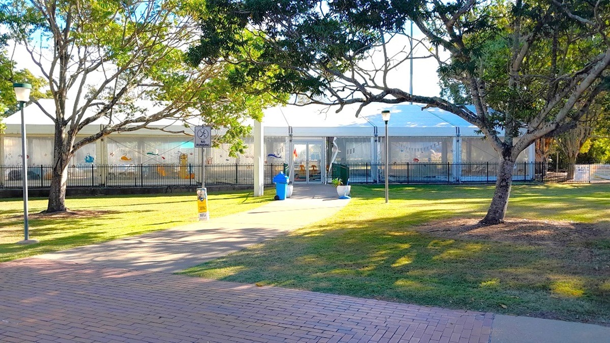 This is an exterior view of the indoor heated pool at Lawnton Aquatic Centre. There is a walkway with a "no running" sign in between the two large trees in front of the main entrance. In addition to two large trees in front of the main entrance, there are also grassed landscape areas. As you walk on the pathway, you will pass by two white pots with plants, a blue trash bin and a black low fence on both sides. A set of double glass doors serve as the main entrance of the indoor pool.