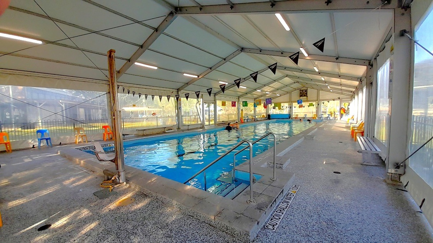 The indoor pool is visible from the inside and has a large white permanent marquee with transparent retractable awning walls, strings of banners with vibrant pennants strung between the ceiling and the blue water surface, a silver access ramp on both sides, colorful monobloc chairs, long benches, and numerous reminders written on the stone flooring and pool sides about the water level.