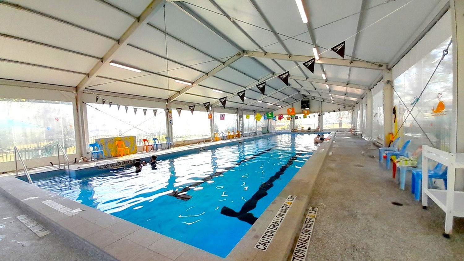 The access ramp is on the left side of the 25-meter pool, where banners with brightly coloured pennants are strung together and hung between the ceiling and the water's surface. Here, colorful plastic chairs are arranged all around the pool, which is surrounded by retractable transparent awning walls that let in natural light. The base of the pool prominently displays the pool's black lane floor markings.