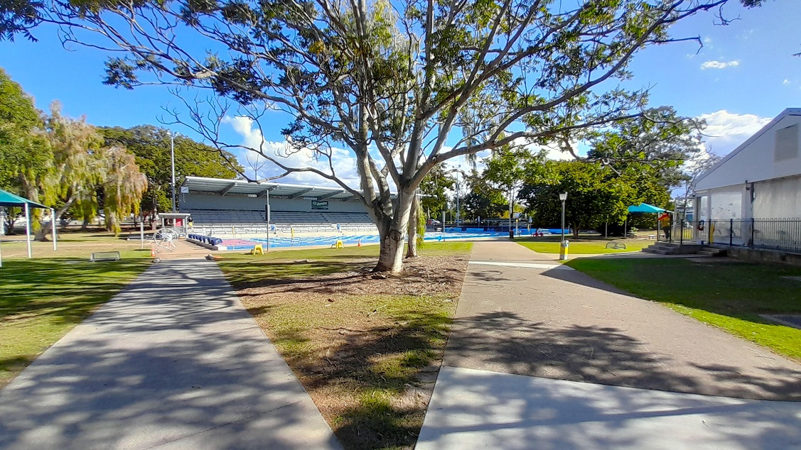 This is a distant view of the outdoor pool, the grandstand and the large trees around it. Two flat concrete pathways have been laid out around the area, and the building with the indoor pool can be seen on the right. The tree in the middle of the outdoor space serves as the focal point. The tree is situated inside a triangular grassed landscape that can be used for picnics.