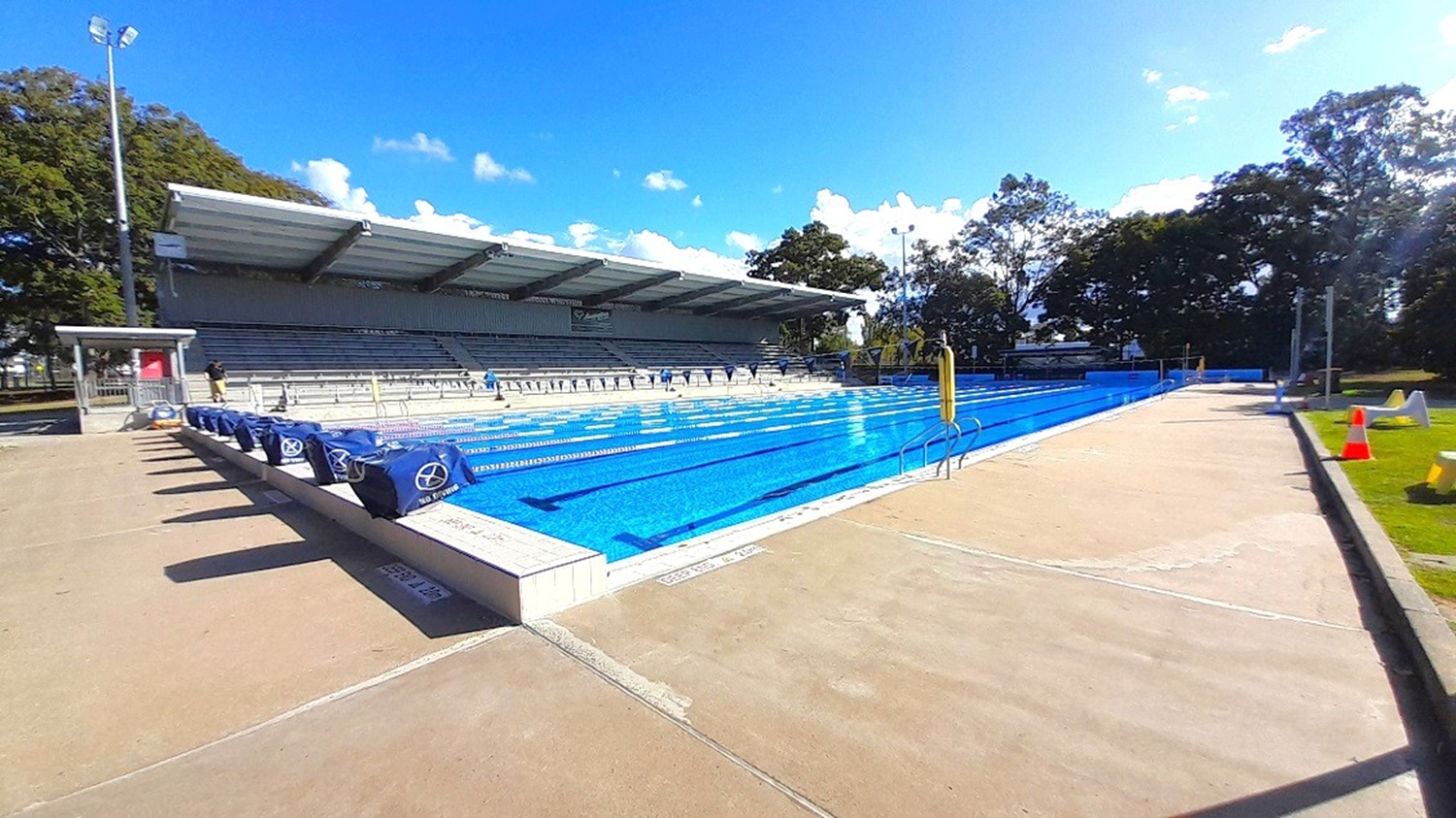 The 50-meter outdoor pool with grey grandstand is the focal point of this view. Accessible ramps are placed on either side of the pool, and "No Diving" signs are lined up along the pool coping. The entire area is shaded by a thick canopy of large trees, and immediately to the left are cemented pool side flooring, which also partially reveals the grassy area where pool chairs are available.