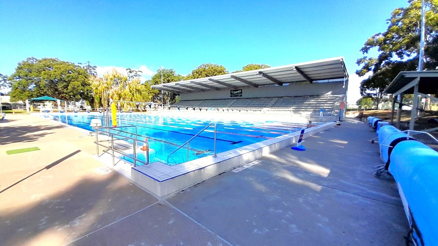 An up-close view of the right pool coping of the outdoor pool and the side of the grey grandstand. The pool side has a grey-brown flooring and the facility is completely shaded by the thick canopy of the big trees surrounding the grandstand. Banners with black pennants are connected by strings attached to the standing silver poles on both sides.