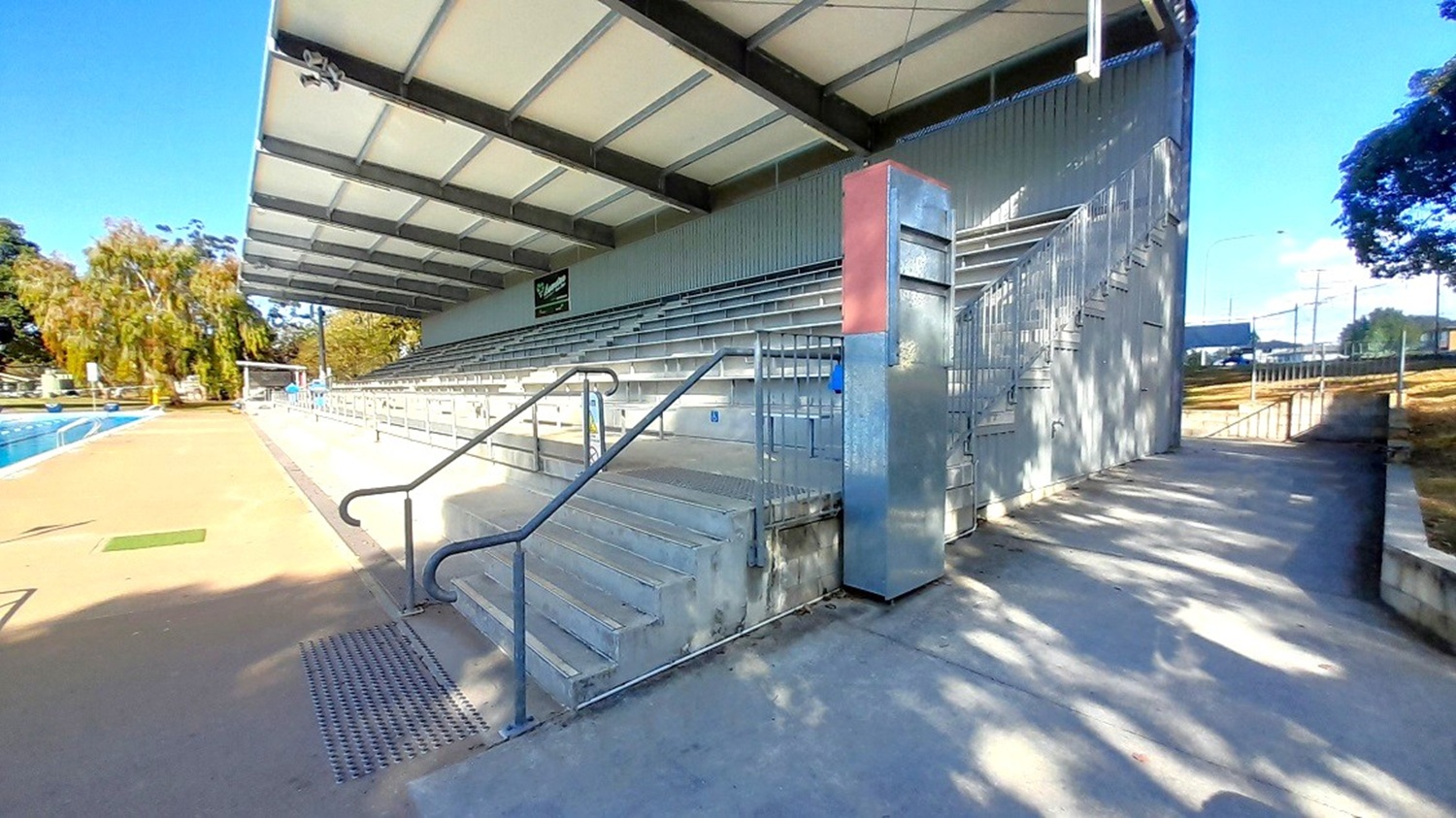 This image shows the side view of the grey-themed grandstand facing the outdoor pool. Its ceiling is white, and its ceiling supports are showing shades of light and dark grey color. The grandstand provides covered seating for hundreds of people, including disability access. A pathway with grey flooring on the left leads to stairs going to the other parts of the center.