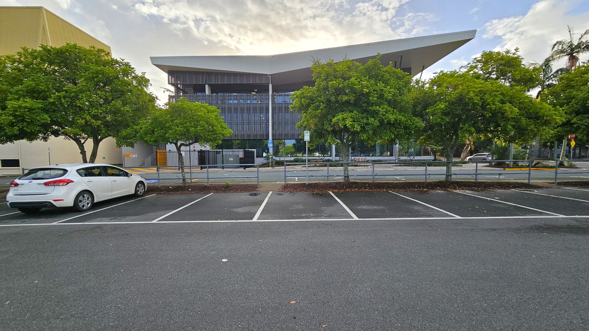A white car is parked on the left side of an empty parking lot. In the background, there are several green trees lining the street. Behind the trees, a modern building with a distinctive angular roof and glass facade is visible. The ground is asphalt, and there are white parking lines. A yellow building is partially visible on the far left.