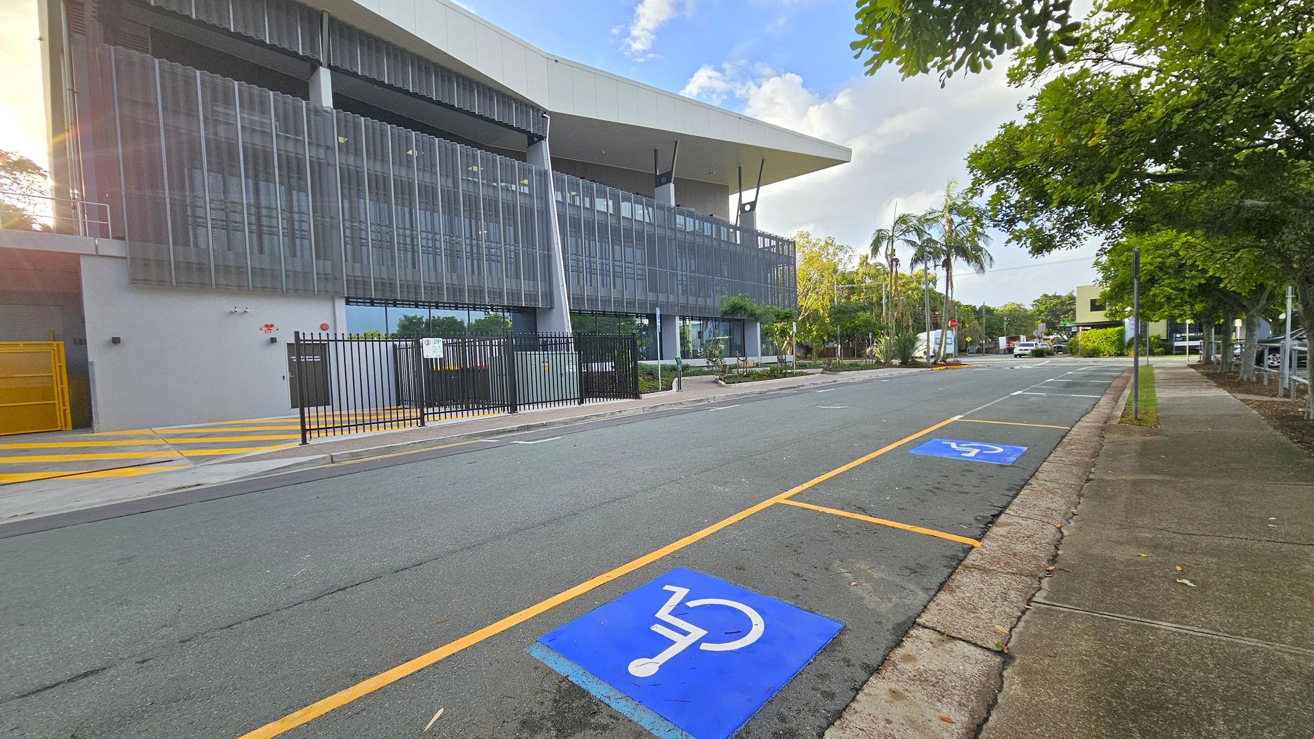 A modern building with a large overhanging roof and vertical metal slats on the left. In front, there is a black metal fence and a yellow ramp. The road has two blue disabled parking spaces marked with white symbols. On the right, a footpath runs alongside the road, bordered by grass and trees, including palm trees. The background features more trees and a few parked vehicles.