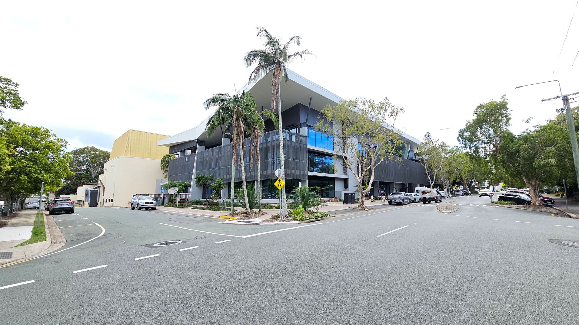 A modern building with a geometric design is situated at a street corner. It features large glass windows and a dark facade. Palm trees and other greenery are planted around the building. On the left, a yellow structure is visible, and several cars are parked along the street. The road is wide, with clear lane markings, and trees line the pavement on both sides. The sky is overexposed, making it appear white.