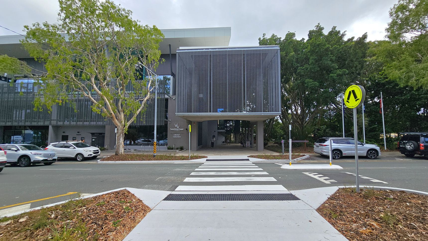 A modern building with a large, grid-like facade is in the centre, with a covered walkway beneath. To the left, a tree with light green leaves stands near parked cars. On the right, a yellow pedestrian crossing sign is visible, with more parked cars and dense trees in the background. A zebra crossing leads towards the building from the foreground, bordered by a landscaped area with mulch and sparse vegetation.